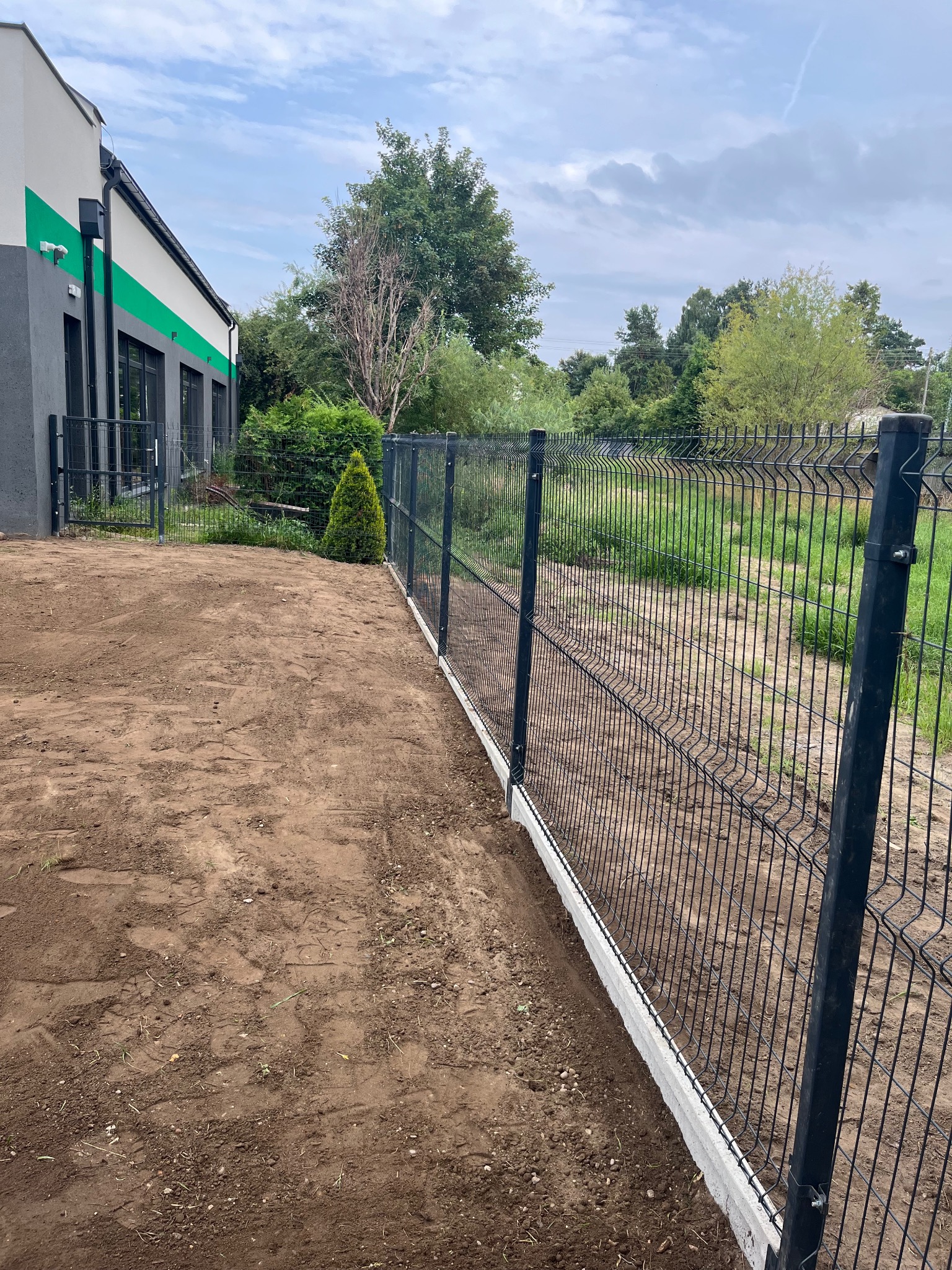 Nowoczesne ogrodzenie panelowe z podmurówką betonową, widok z boku na tle budynku i zieleni. Czarna siatka ogrodzeniowa, świeżo obsypana ziemią.