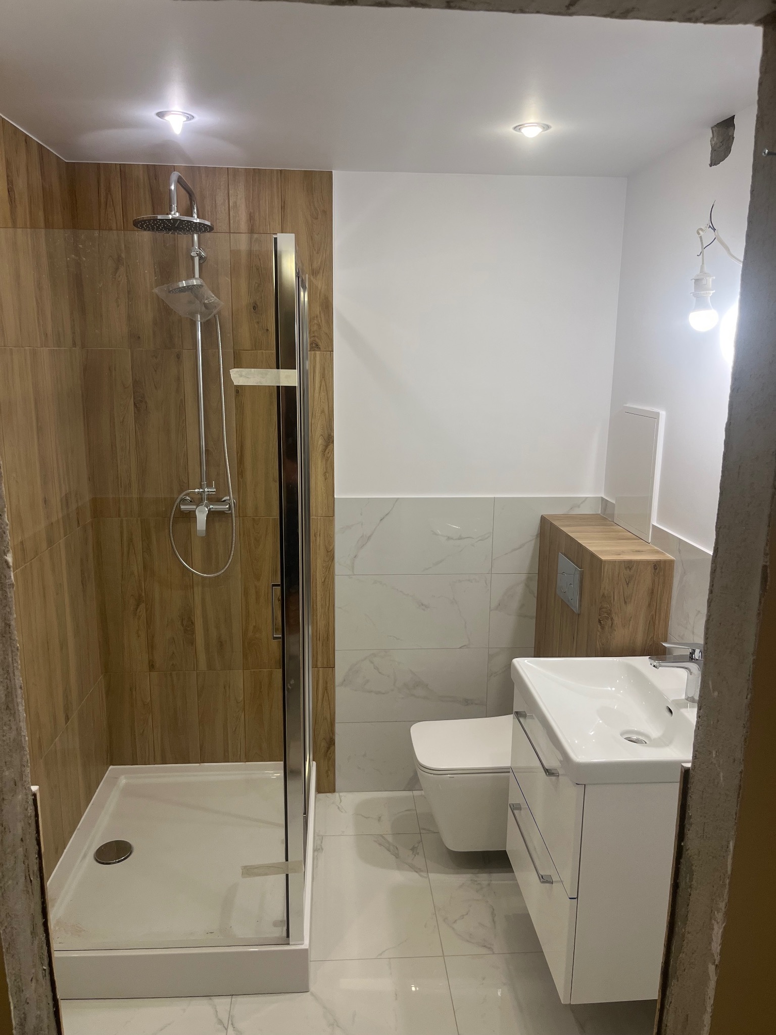 Nowoczesna łazienka po remoncie: kabina prysznicowa z drewnopodobnymi płytkami, biała umywalka z szafką i toaleta. Marmurowe płytki na ścianach i podłodze.