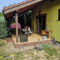 Maciej Rokita - Przerobiony taras na salon z żółtą elewacją, wyłożony płytkami, z drewnianym zadaszeniem i stołem, przy którym siedzą ludzie. Widoczne narzędzia ogrodowe.