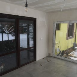 Maciej Rokita - Wnętrze w trakcie wykańczania: ułożone szare płytki, ściany przygotowane pod malowanie, widoczne okna z widokiem na zimowy krajobraz i choinkę. Częściowo wykończony sufit z płyt g-k.