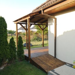 Maciej Rokita - Drewniana altana przylegająca do budynku z brązowym dachem i rynną. Widoczny fragment tarasu z desek, trawnik i tuje. Ujęcie z poziomu trawnika, wczesne popołudnie.