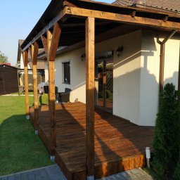 Maciej Rokita - Drewniana, zadaszona weranda przy domu z brązową dachówką, oświetlona popołudniowym słońcem. Widoczne meble ogrodowe i zadbany trawnik.