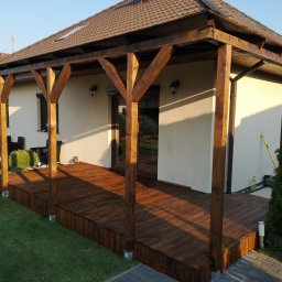 Maciej Rokita - Zadaszony taras z drewnianą podłogą i konstrukcją wsporczą, przylegający do jasnego budynku z oknami i zielonym trawnikiem. Widoczne detale wykończenia i oświetlenie.