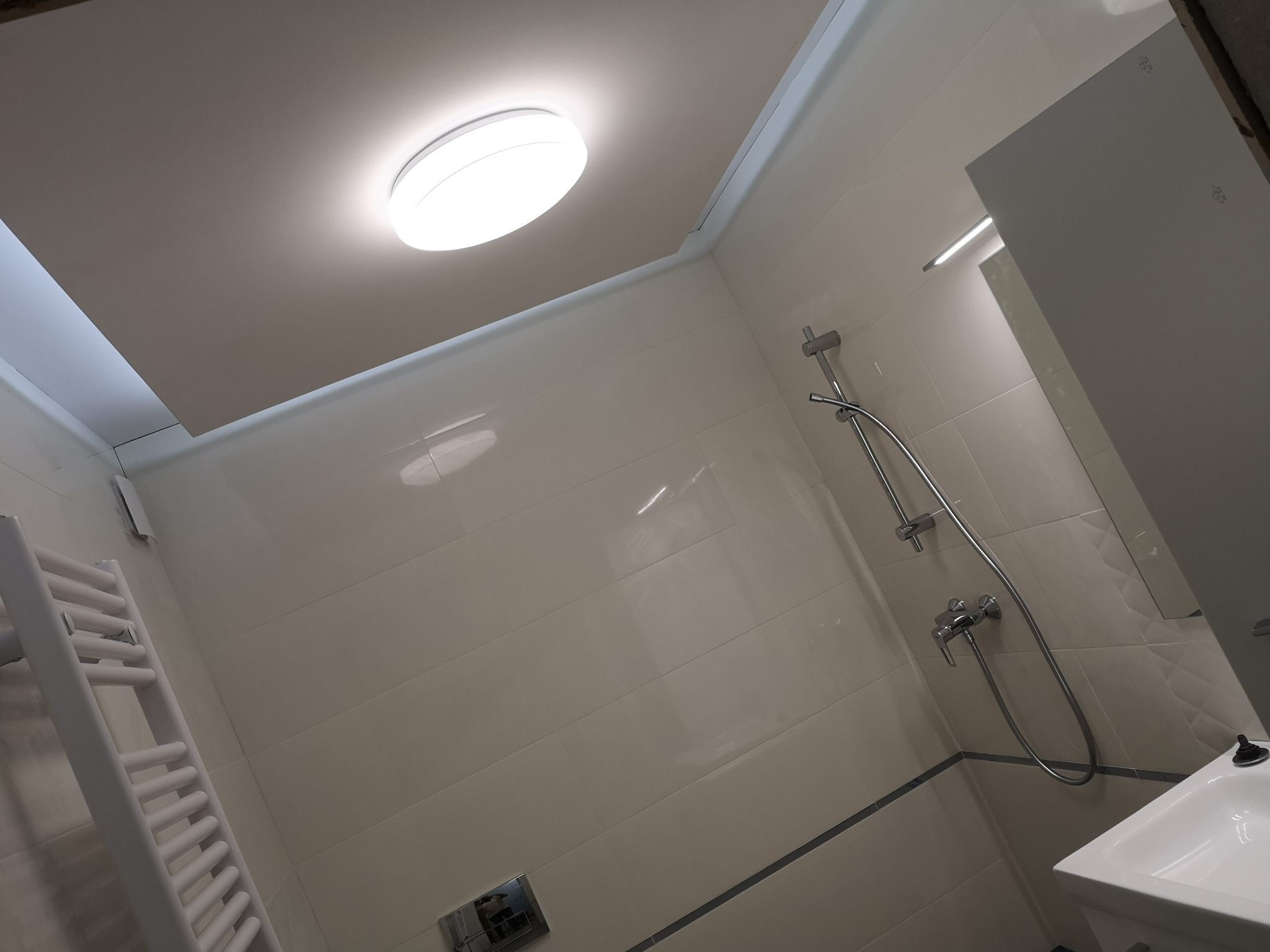 Jasna łazienka po remoncie z beżowymi płytkami, grzejnikiem drabinkowym i kabiną prysznicową. Oświetlenie sufitowe LED. Minimalistyczny design.