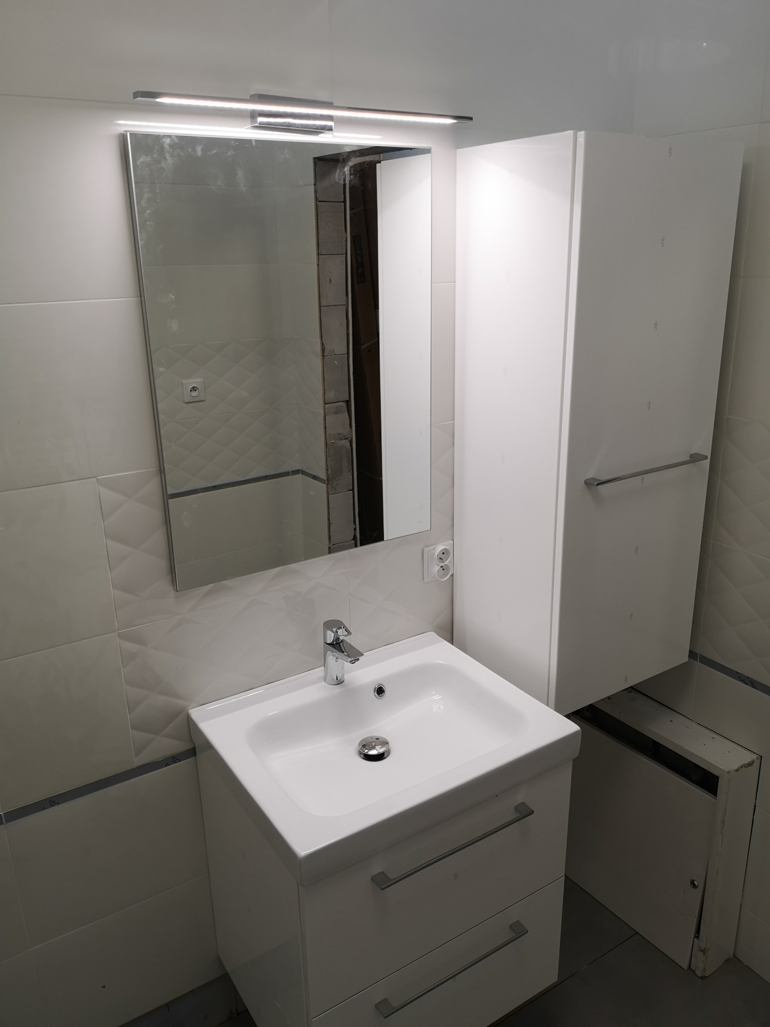 Biała umywalka z szafką i lustrem z oświetleniem w łazience wykończonej jasnymi płytkami. Widoczna szafka wisząca i fragment instalacji w odbiciu lustra.