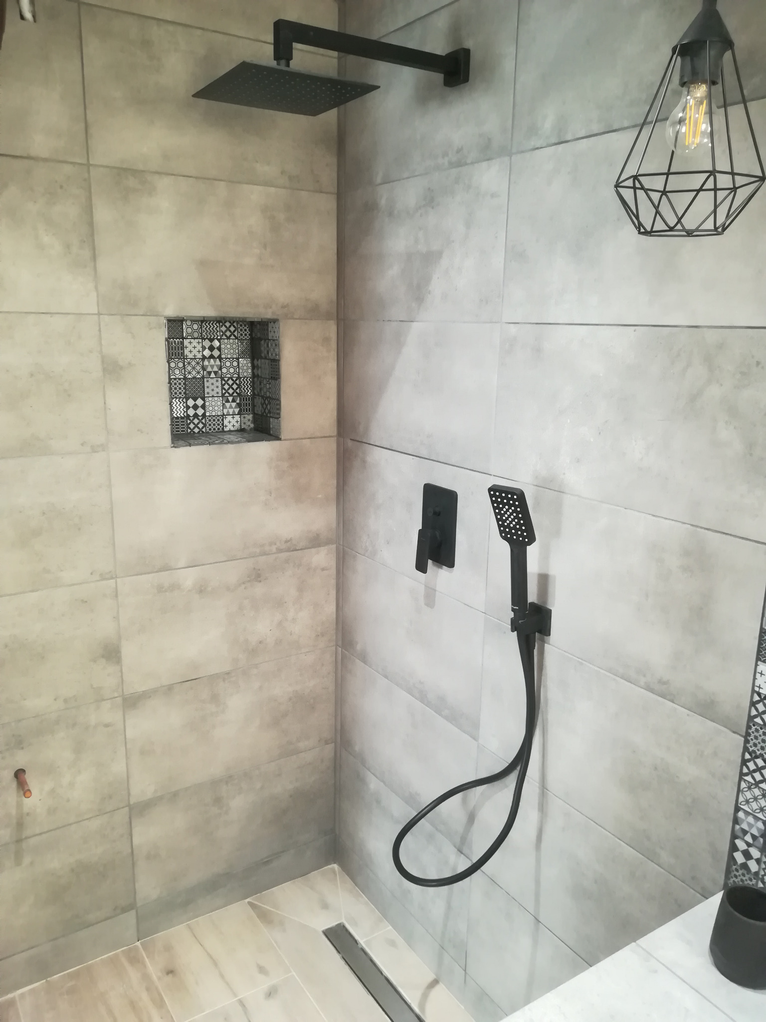 Nowoczesna łazienka z szarymi płytkami imitującymi beton, czarną armaturą i dekoracyjną wnęką z mozaiką. Widoczny odpływ liniowy i designerska lampa.