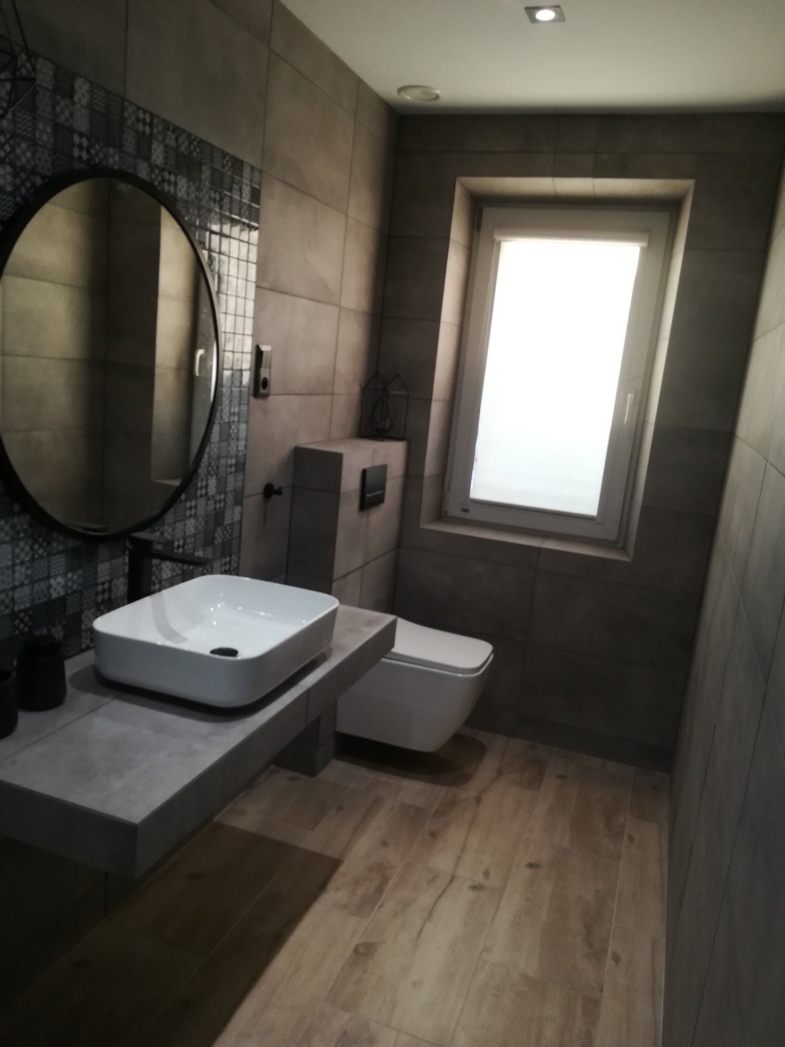 Nowoczesna łazienka w szarościach: okrągłe lustro, kwadratowa umywalka, podwieszana toaleta i drewnopodobne płytki podłogowe. Minimalistyczny design z akcentem na geometryczne formy.