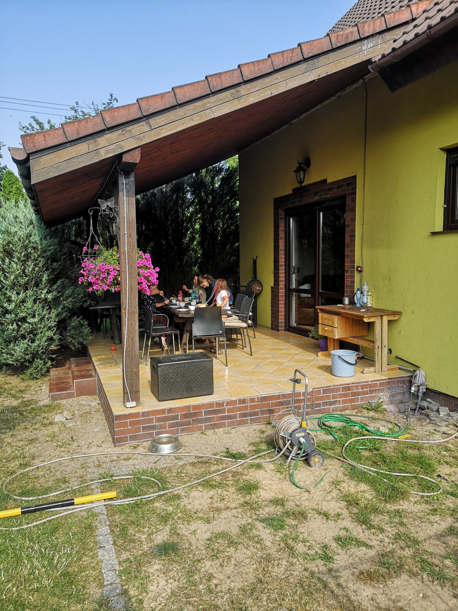 Przerobiony taras na salon z żółtą elewacją, wyłożony płytkami, z drewnianym zadaszeniem i stołem, przy którym siedzą ludzie. Widoczne narzędzia ogrodowe.