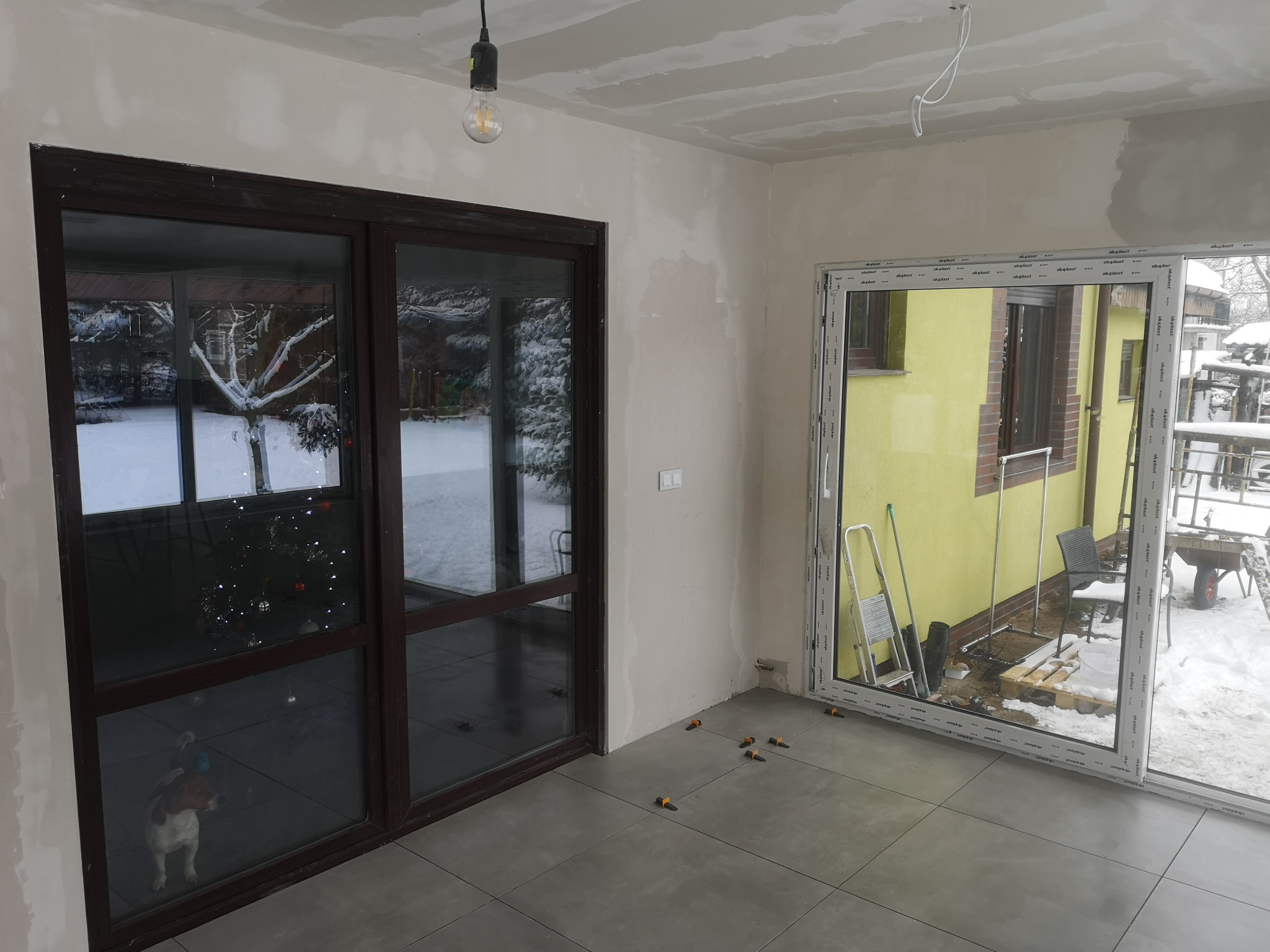 Wnętrze w trakcie wykańczania: ułożone szare płytki, ściany przygotowane pod malowanie, widoczne okna z widokiem na zimowy krajobraz i choinkę. Częściowo wykończony sufit z płyt g-k.