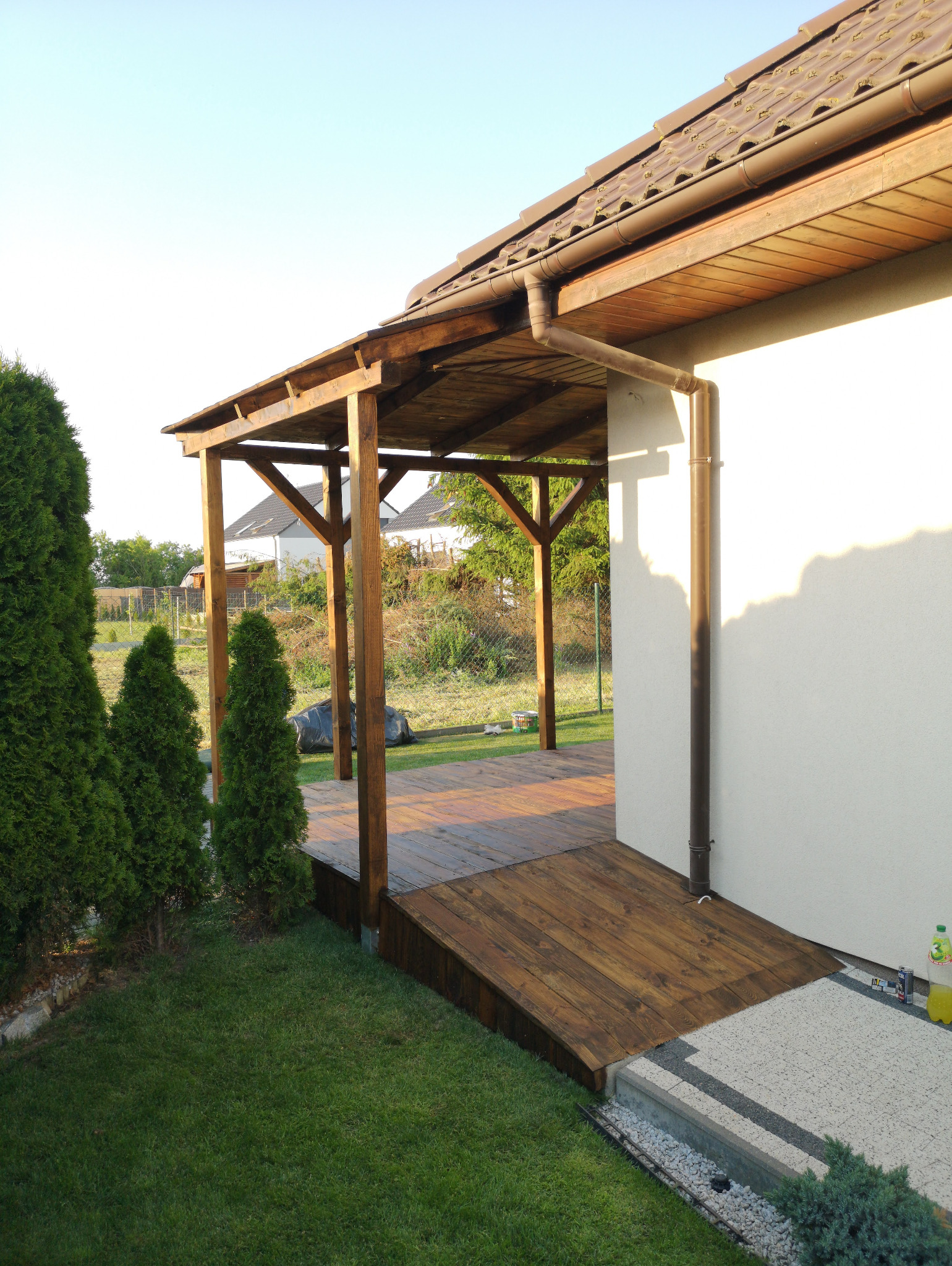 Drewniana altana przylegająca do budynku z brązowym dachem i rynną. Widoczny fragment tarasu z desek, trawnik i tuje. Ujęcie z poziomu trawnika, wczesne popołudnie.
