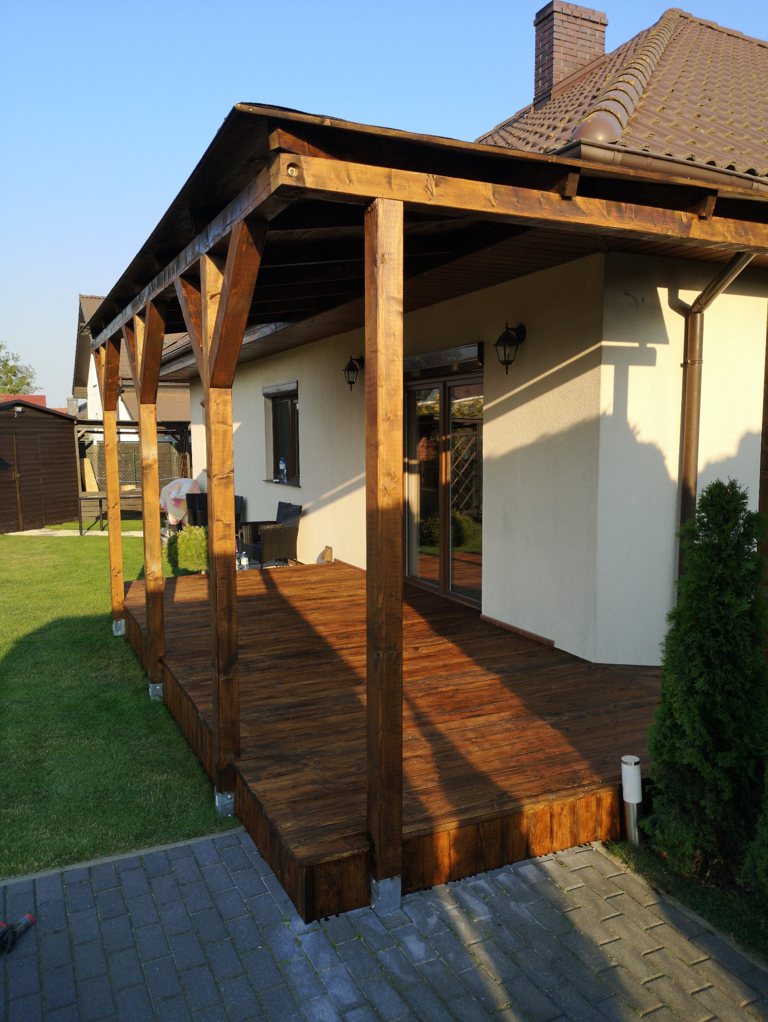 Drewniana, zadaszona weranda przy domu z brązową dachówką, oświetlona popołudniowym słońcem. Widoczne meble ogrodowe i zadbany trawnik.