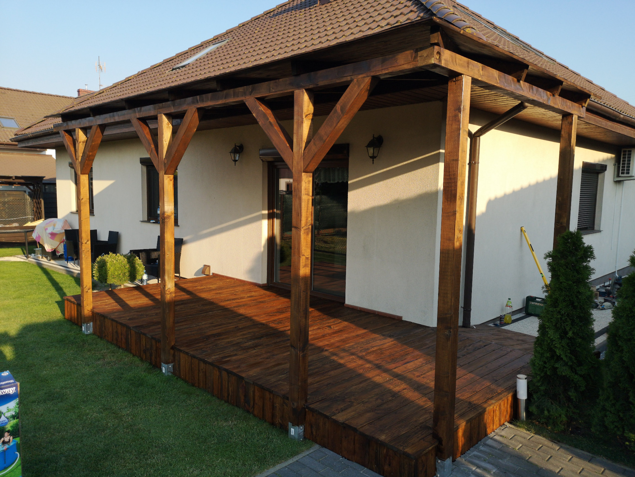 Zadaszony taras z drewnianą podłogą i konstrukcją wsporczą, przylegający do jasnego budynku z oknami i zielonym trawnikiem. Widoczne detale wykończenia i oświetlenie.