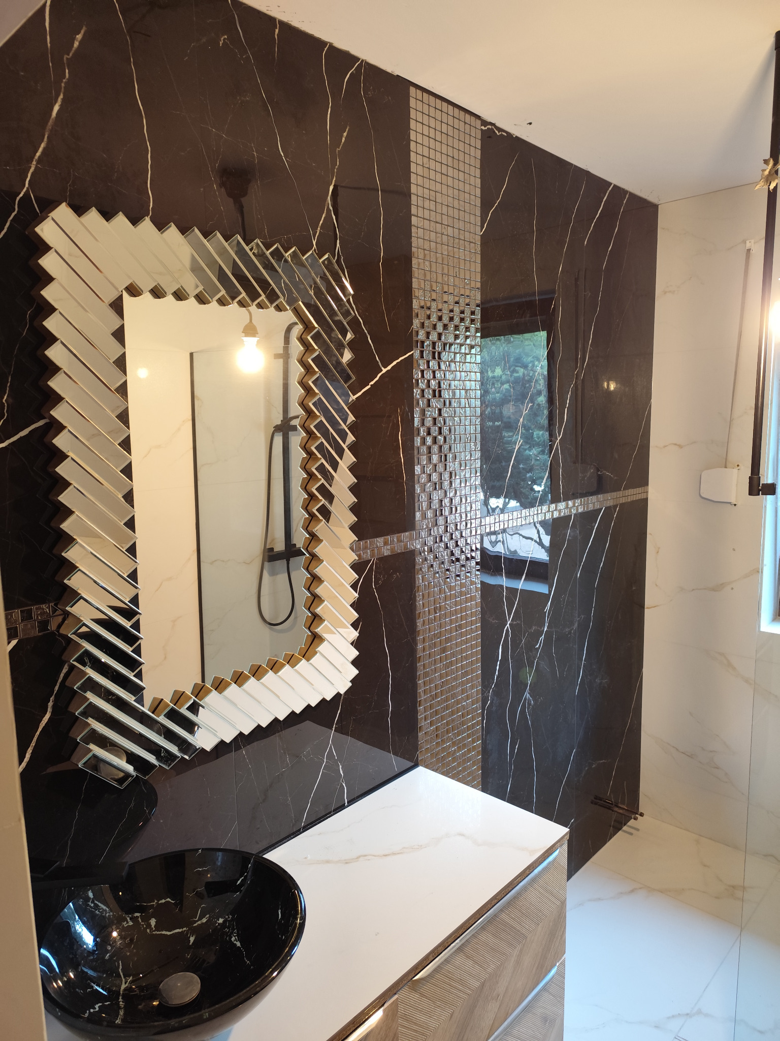 Nowoczesna łazienka z czarnymi płytkami imitującymi marmur, mozaiką i lustrem w ozdobnej ramie. Umywalka nablatowa w kolorze czarnym.