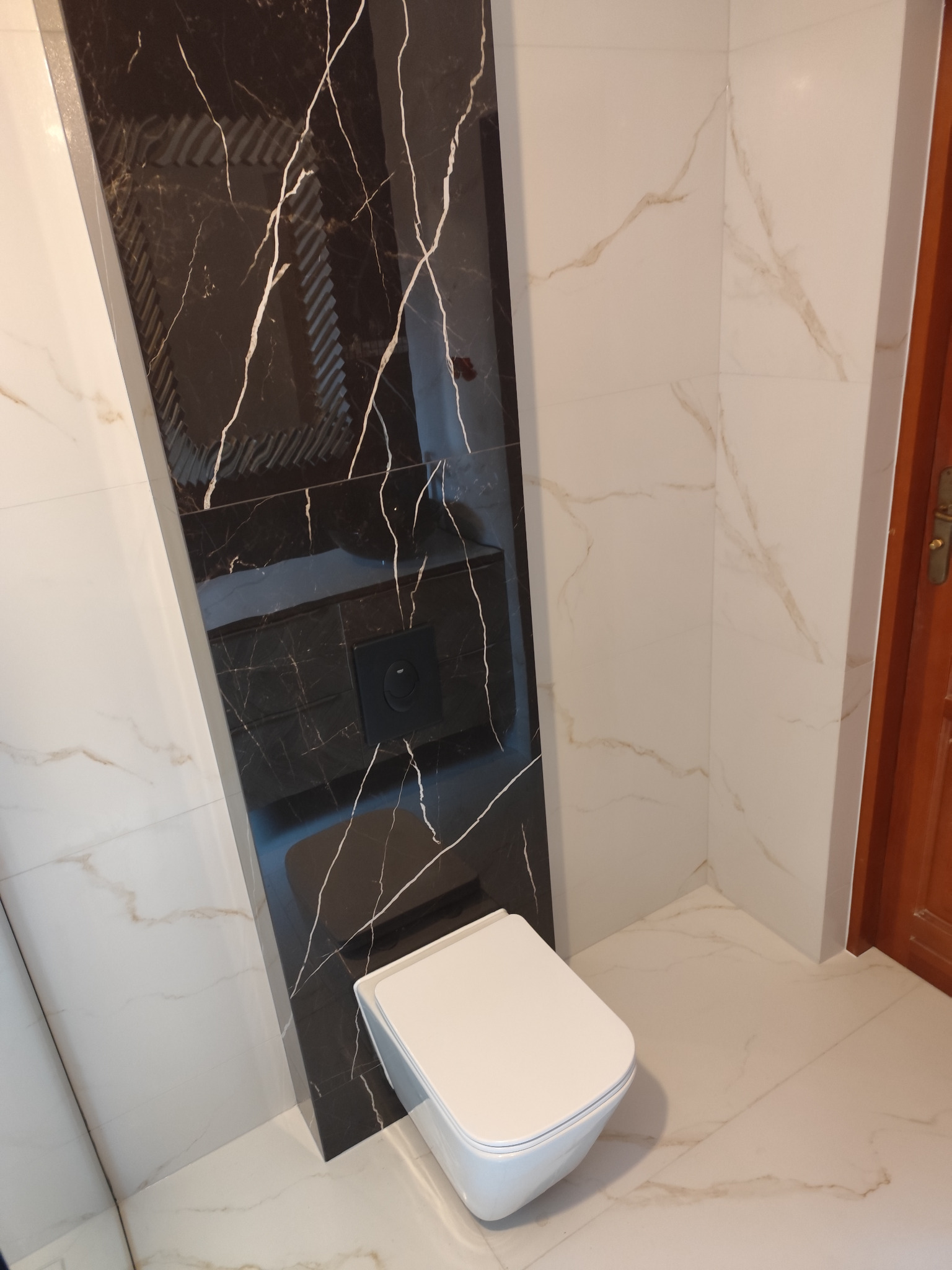 Nowoczesna łazienka z marmurowymi płytkami w odcieniach bieli i czerni, wisząca toaleta, minimalistyczny design, drewniane drzwi w tle.