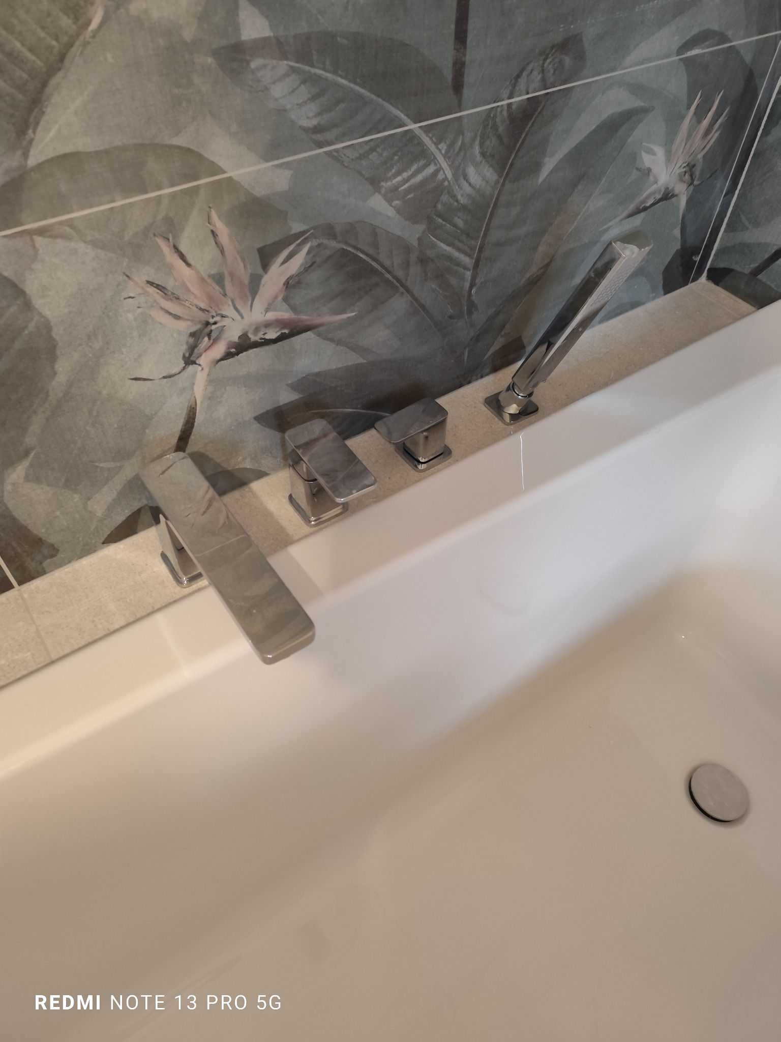 Bateria wannowa z deszczownicą na tle płytek z motywem tropikalnym. Nowoczesna armatura w łazience po remoncie. Minimalistyczny design.