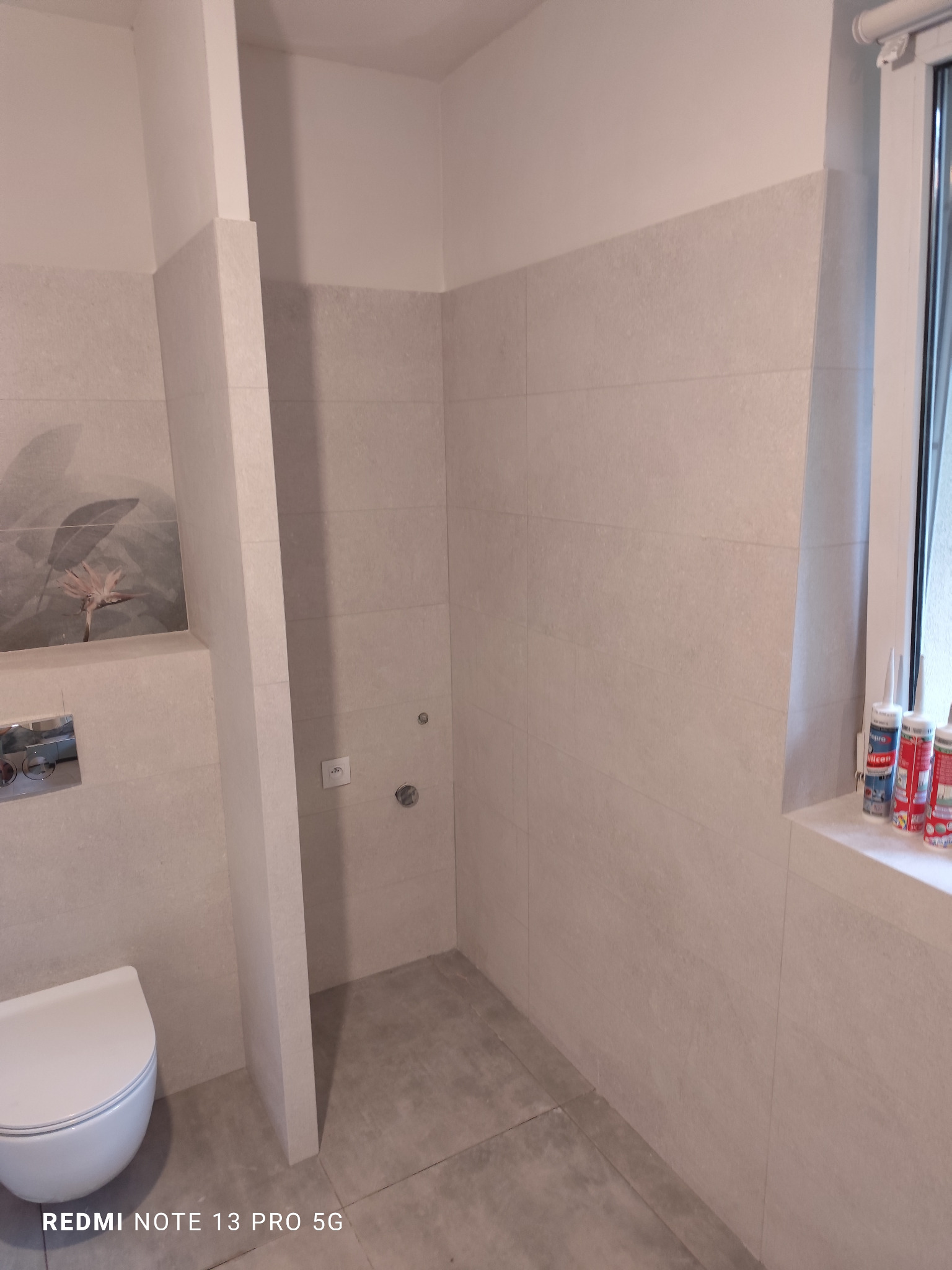 Wykończona łazienka z jasnymi, kwadratowymi płytkami na ścianach i podłodze. Widoczna toaleta, fragment okna z silikonami i dekoracja na ścianie. Minimalistyczny design.