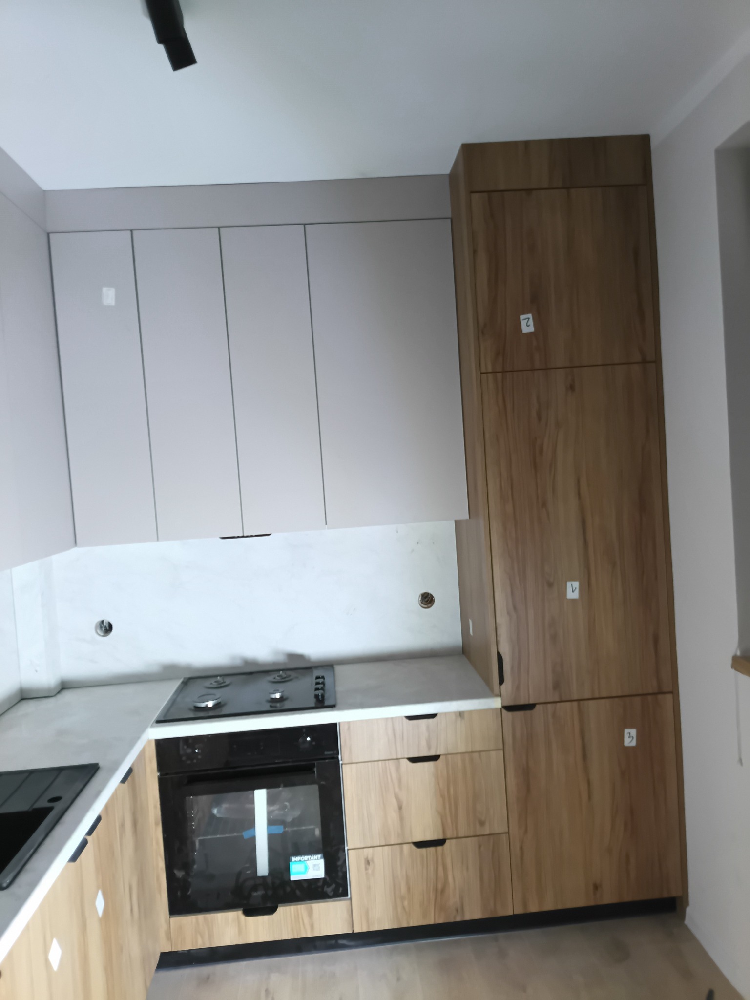 Jasna kuchnia z drewnianymi i szarymi frontami. Widoczny piekarnik, płyta gazowa i wysoka zabudowa. Na frontach naklejone białe karteczki z numerami.