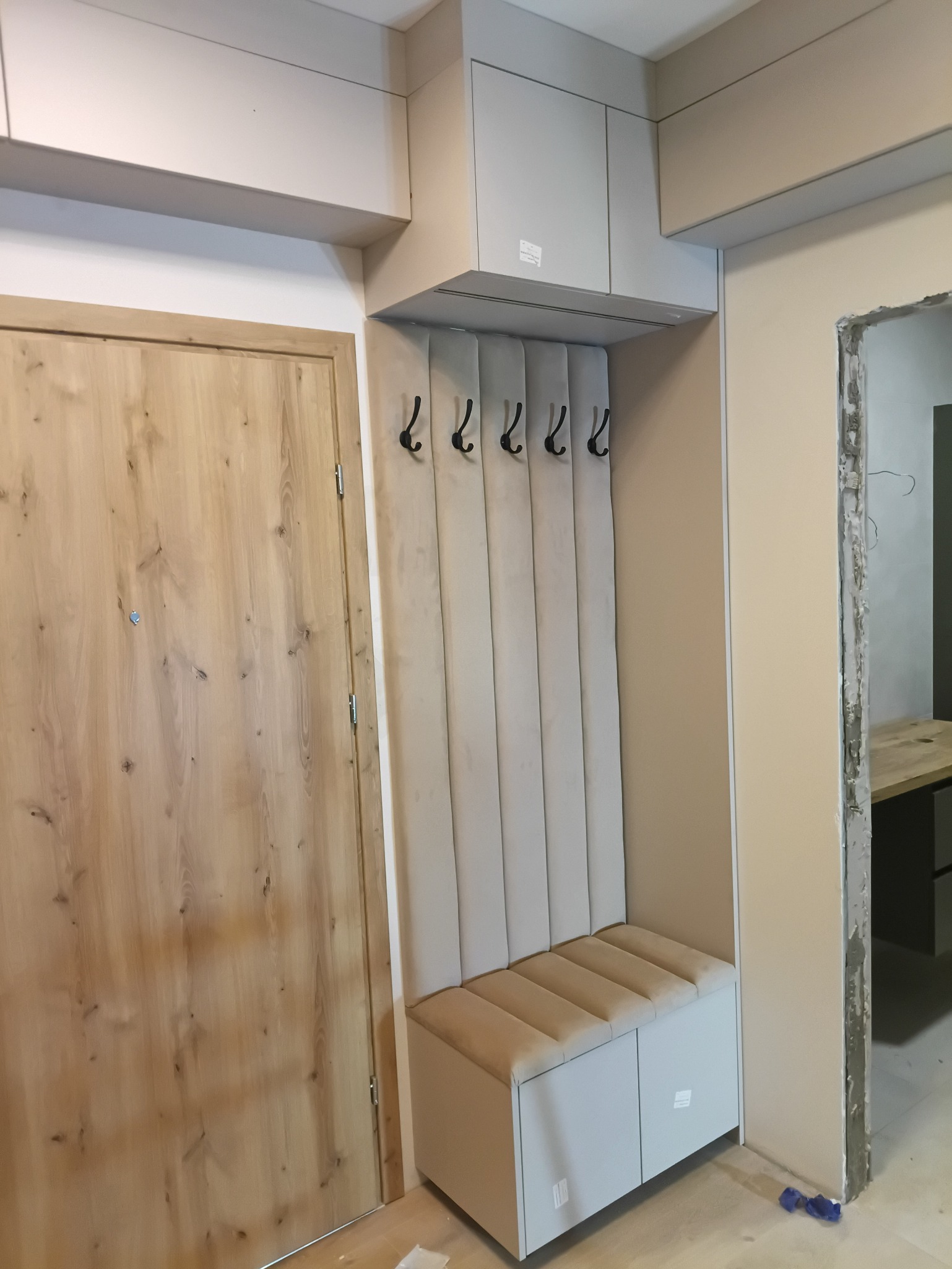 Komplet mebli do przedpokoju w jasnych barwach: szafka z siedziskiem, wieszak panelowy z haczykami i szafka wisząca. Drzwi wejściowe z jasnego drewna po lewej.