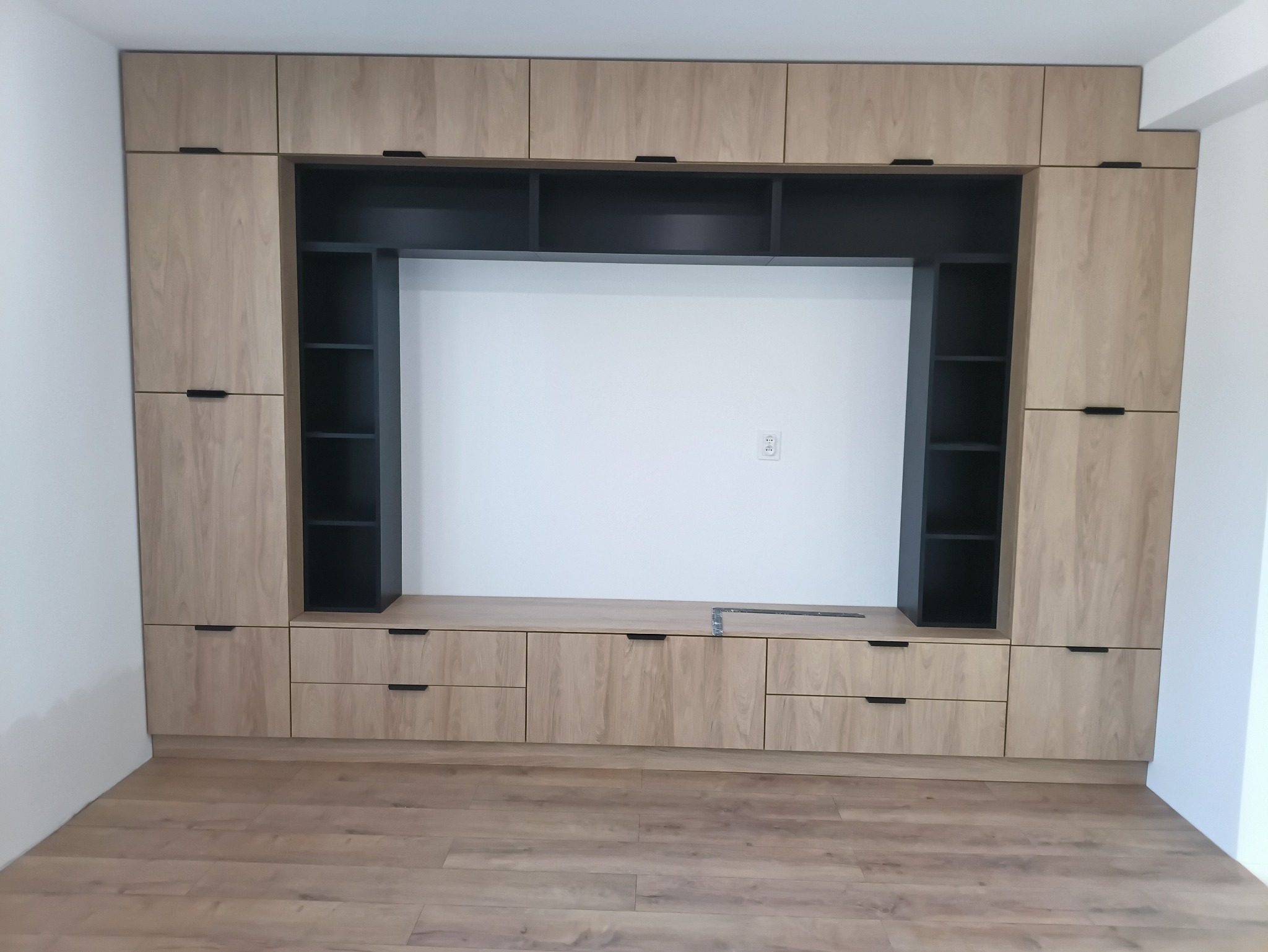 Zabudowa meblowa z jasnego drewna z czarnymi akcentami, obejmująca szafki górne, dolne i otwarte półki. Na blacie leży miarka. Widoczna podłoga z paneli.