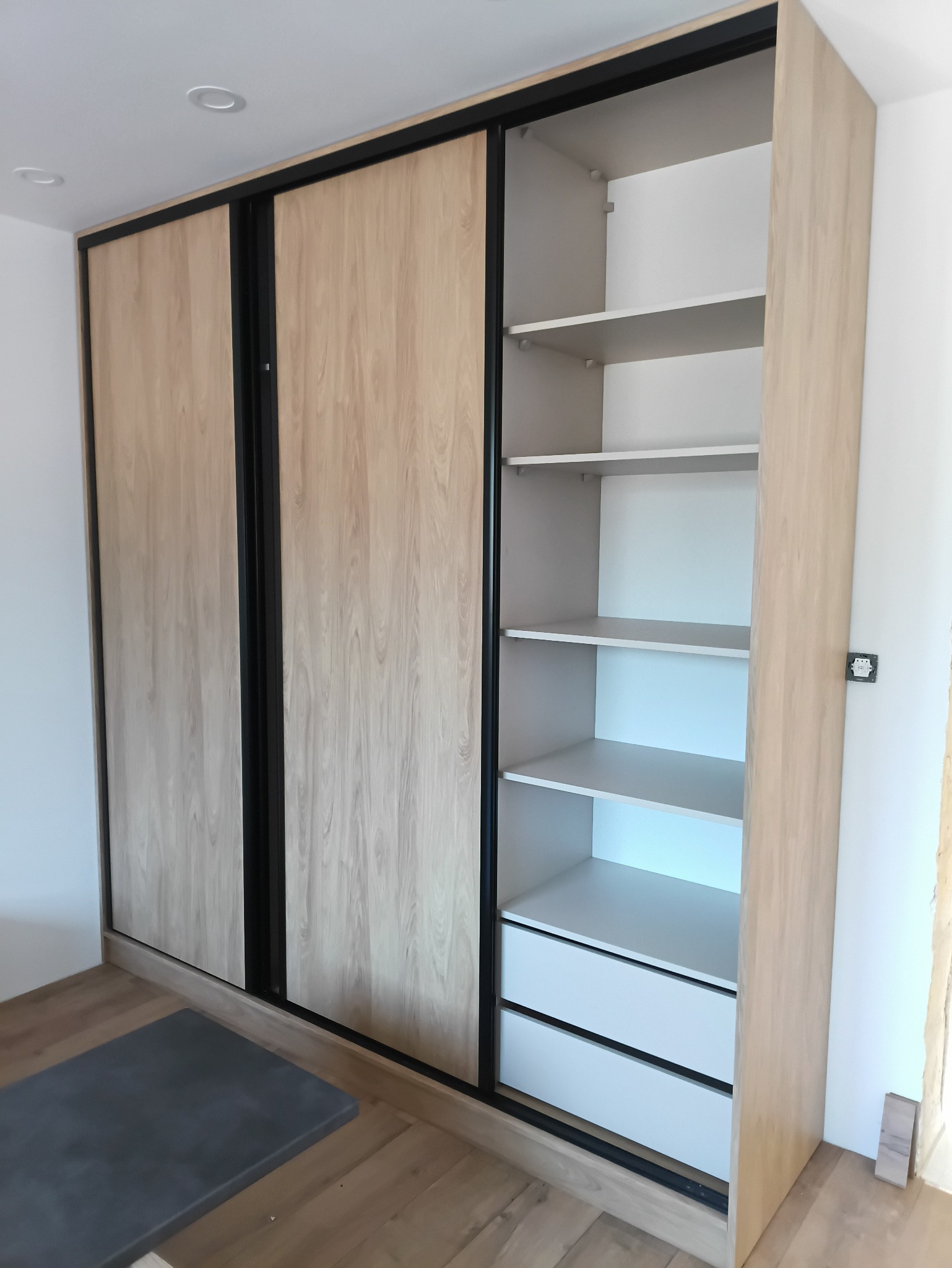 Jasna szafa wnękowa z przesuwnymi drzwiami o wzorze drewna i czarnymi obramowaniami. Widoczne półki i szuflady wewnątrz. Podłoga drewniana, ściany białe.