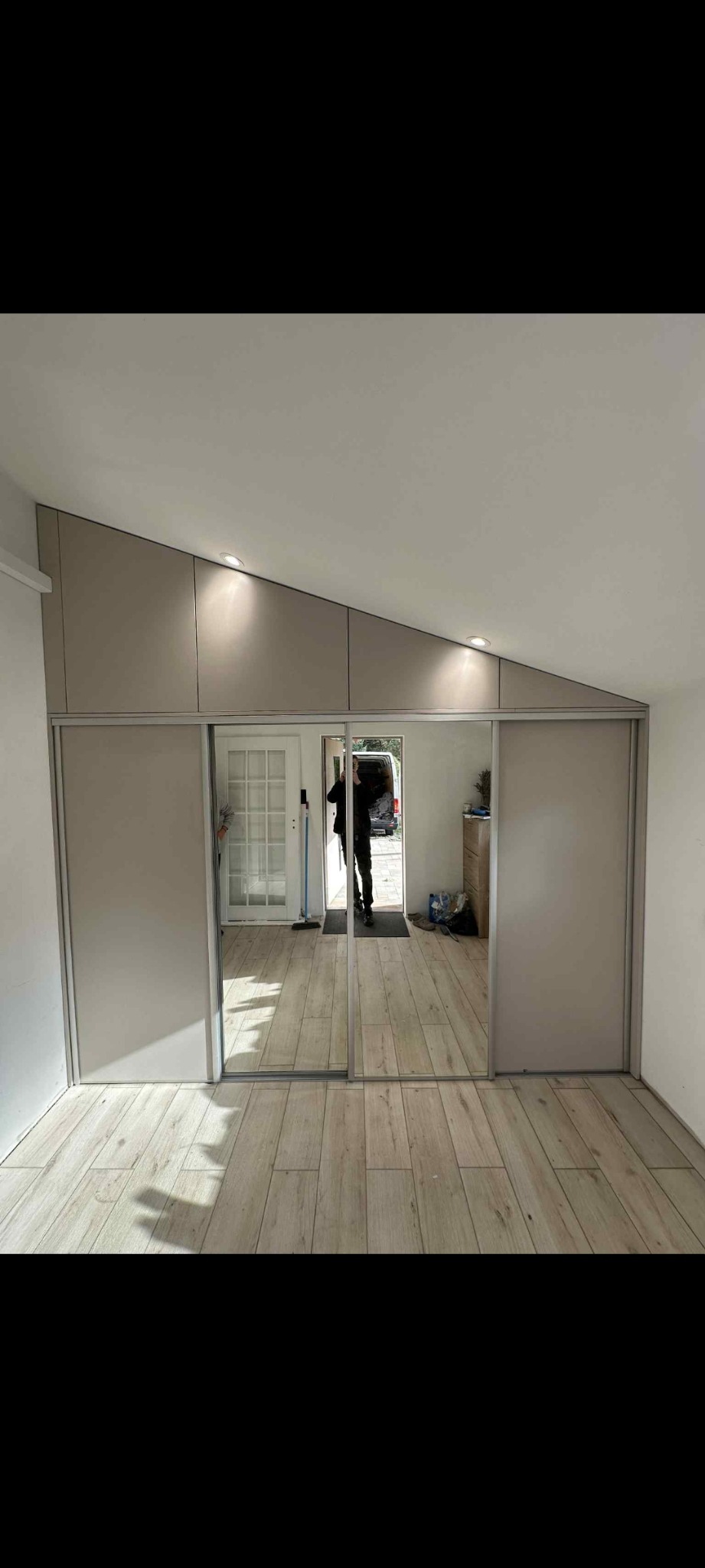 Beżowa szafa wnękowa z przesuwnymi lustrzanymi drzwiami, dopasowana do skosu sufitu. Widoczna podłoga z paneli i odbicie osoby w lustrze.