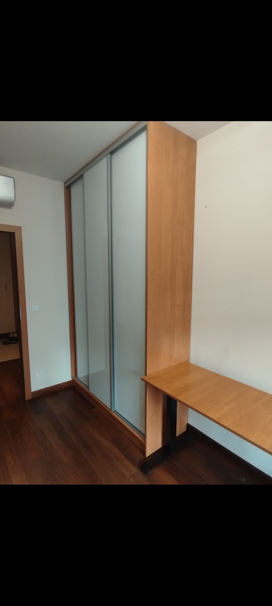 Jasna szafa wnękowa z przesuwnymi drzwiami i biurkiem wbudowanym z boku, na tle drewnianej podłogi i białych ścian. Minimalistyczny design.