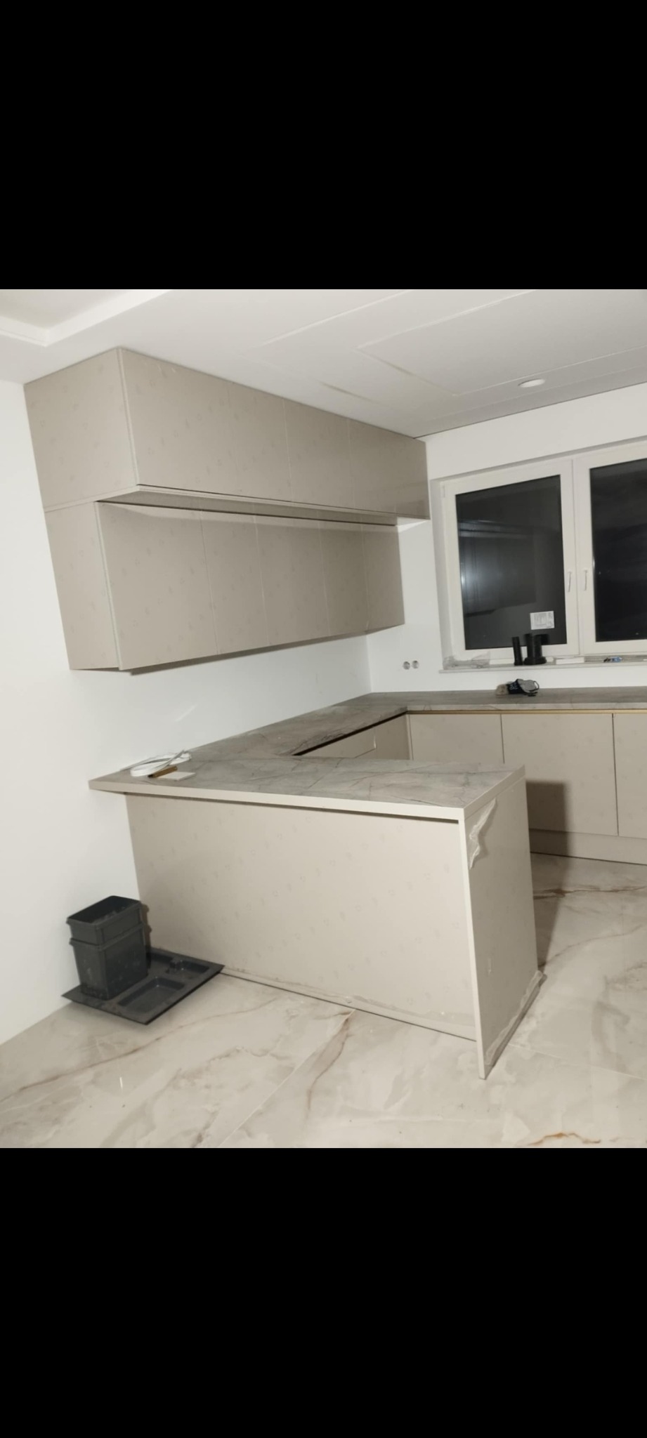 Jasna, nowoczesna kuchnia z wyspą i górnymi szafkami, widok z boku. Na podłodze marmurowe płytki. Widoczne narzędzia i ślady po montażu.