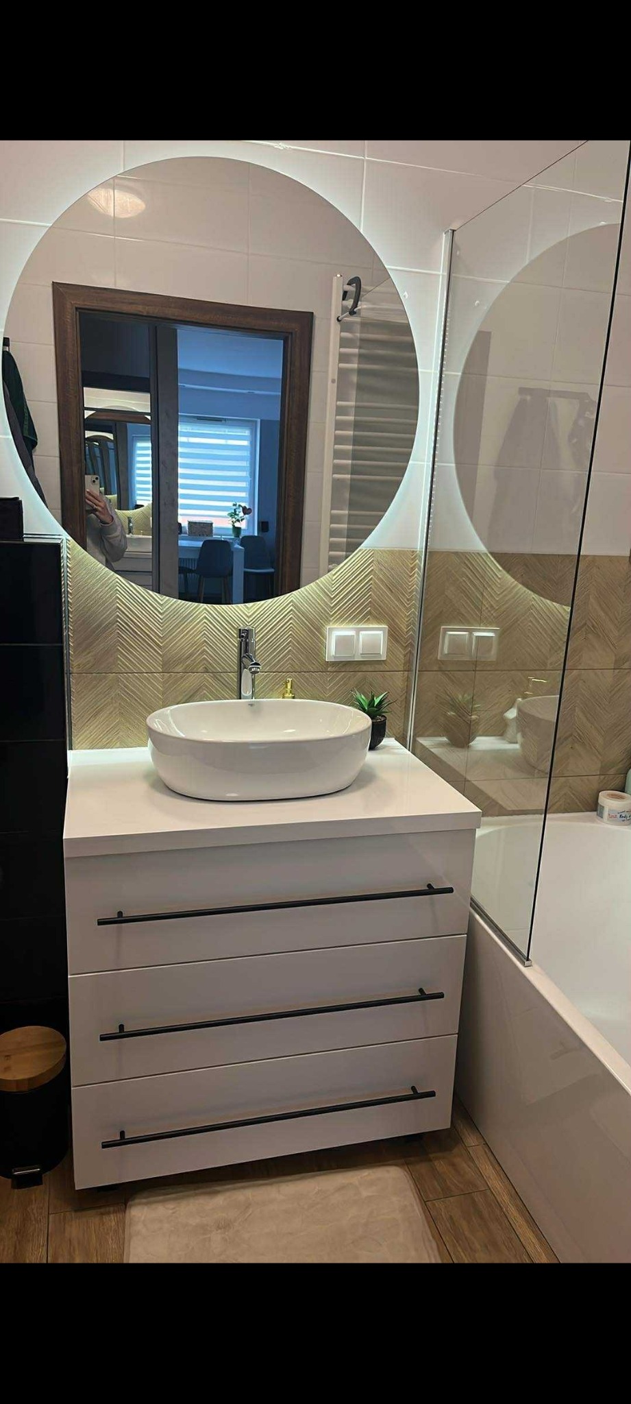 Nowoczesna łazienka z białą szafką pod umywalkę z trzema szufladami i okrągłym lustrem z podświetleniem LED. Na ścianie dekoracyjne płytki w jodełkę.