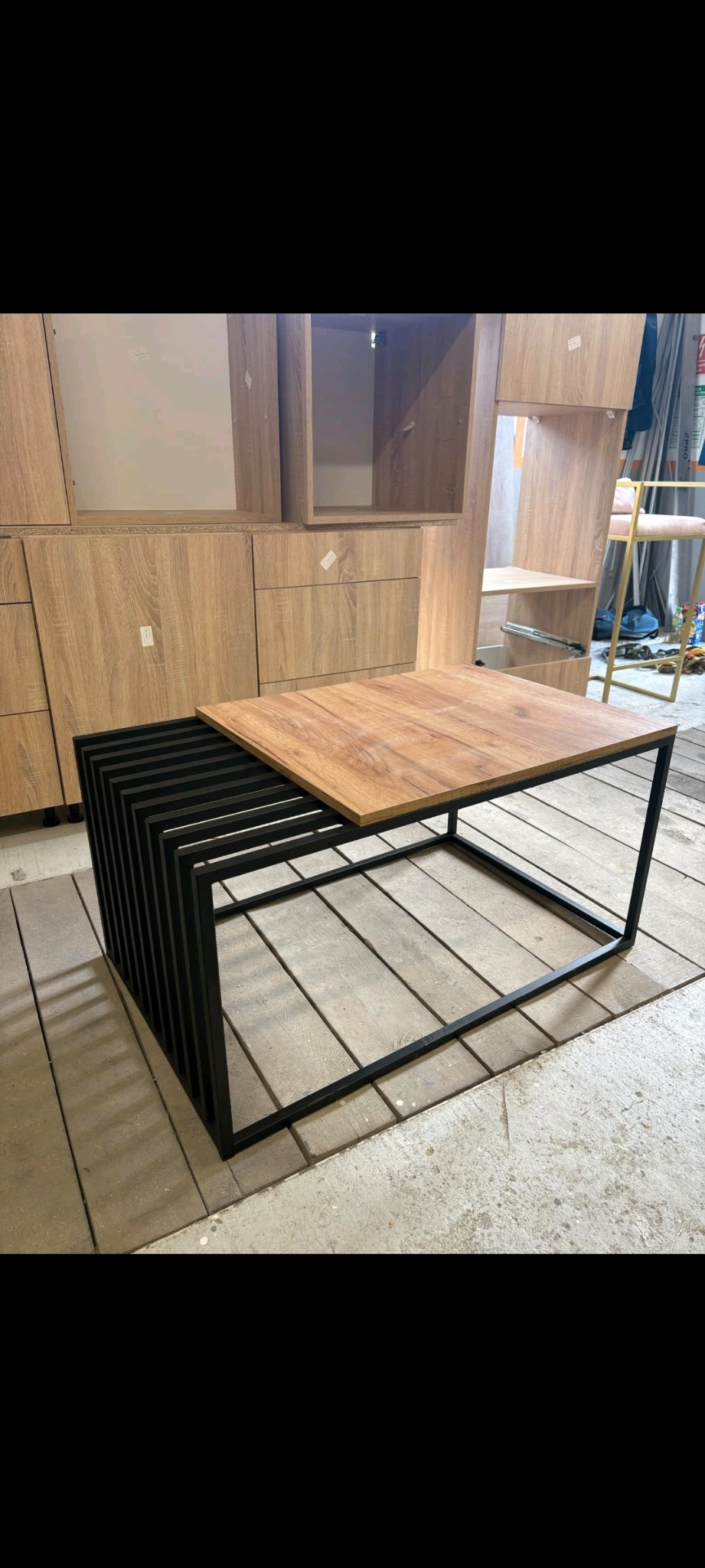 Stylowy stolik kawowy z drewnianym blatem i czarną, metalową podstawą o geometrycznym designie. W tle zabudowa meblowa w odcieniach drewna.