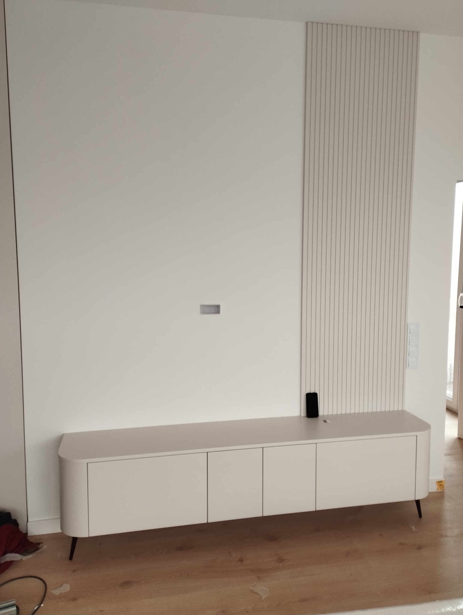 Biała szafka RTV z zaokrąglonymi krawędziami na cienkich nóżkach, na tle ściany z dekoracyjnymi panelami pionowymi. Minimalistyczny design, jasne wnętrze.