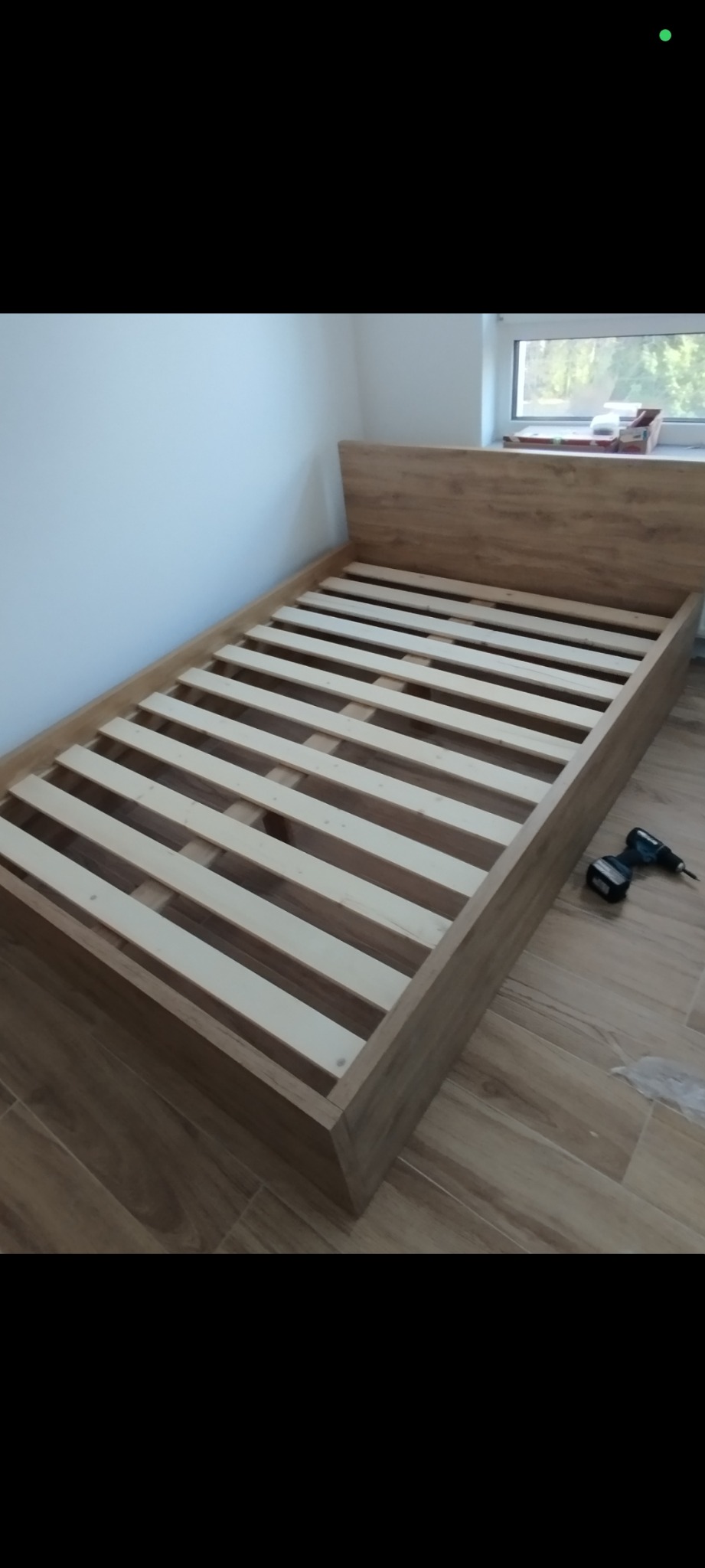 Drewniane łóżko z listwowym stelażem, częściowo zmontowane, z wkrętarką obok, na podłodze imitującej drewno, w jasnym pokoju z oknem.