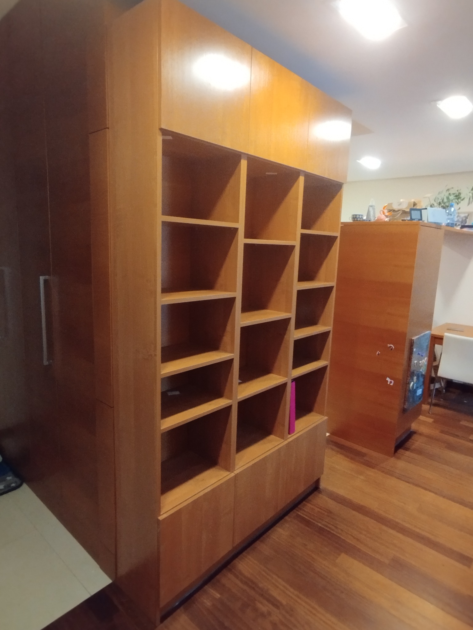 Drewniana szafa wnękowa z otwartymi półkami i zamkniętą częścią, dopasowana do drewnianej podłogi. Widoczne oświetlenie sufitowe i fragment wnętrza.