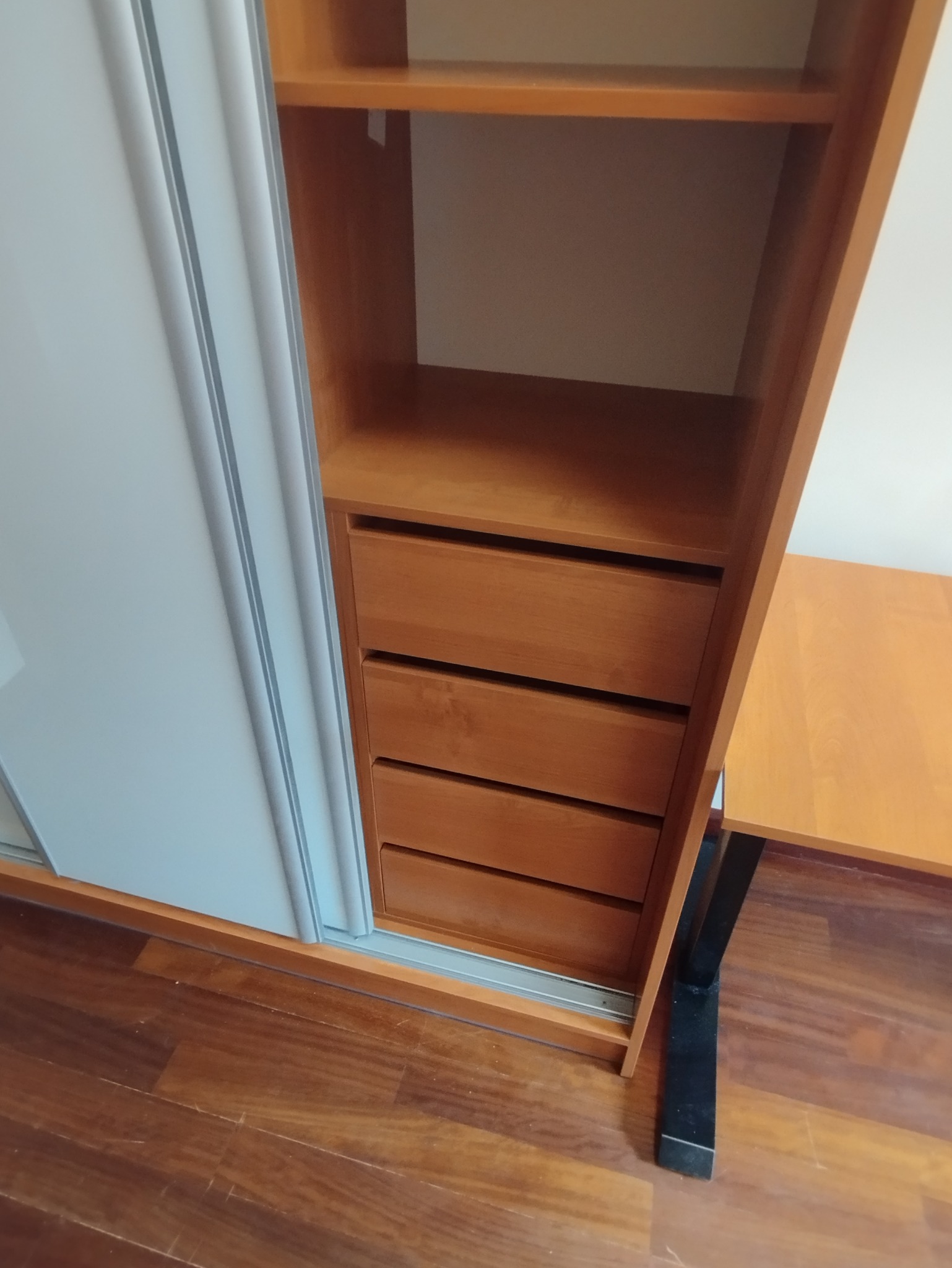 Drewniana szafa z przesuwnymi, białymi drzwiami i szufladami, widoczna na tle parkietu. Ujęcie z bliska, ukazujące detale wykończenia i funkcjonalność mebla.