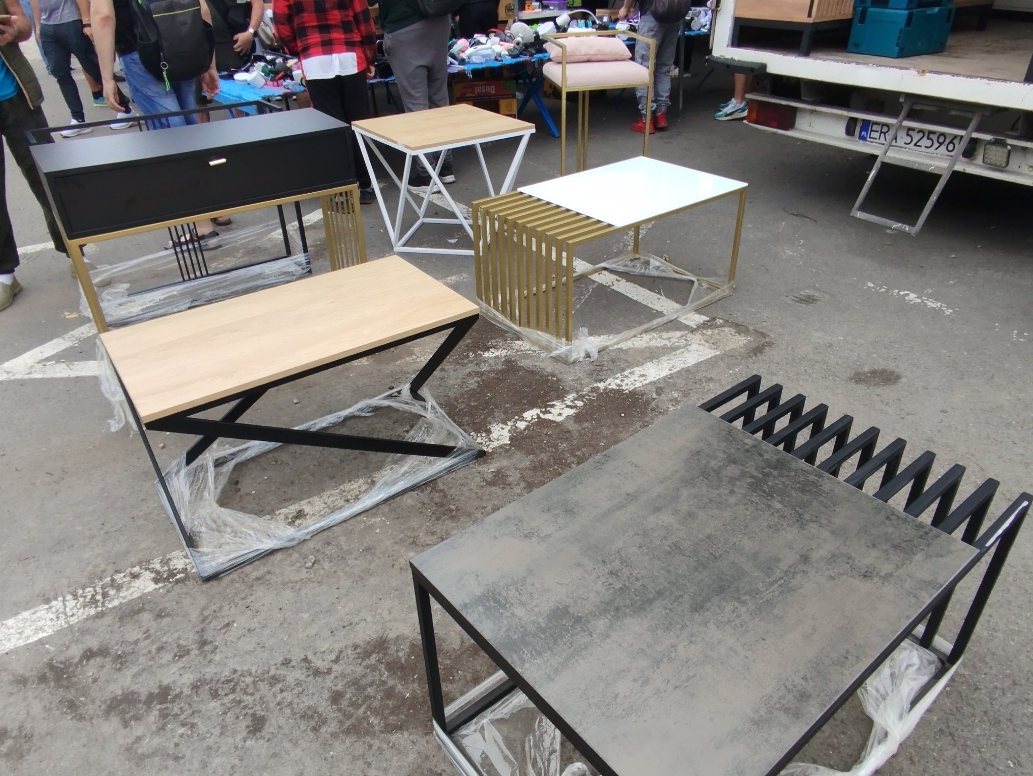 Różnorodne stoliki kawowe z metalowymi nogami i blatami z drewna, szkła i imitacji kamienia, częściowo owinięte folią, ustawione na zewnątrz.