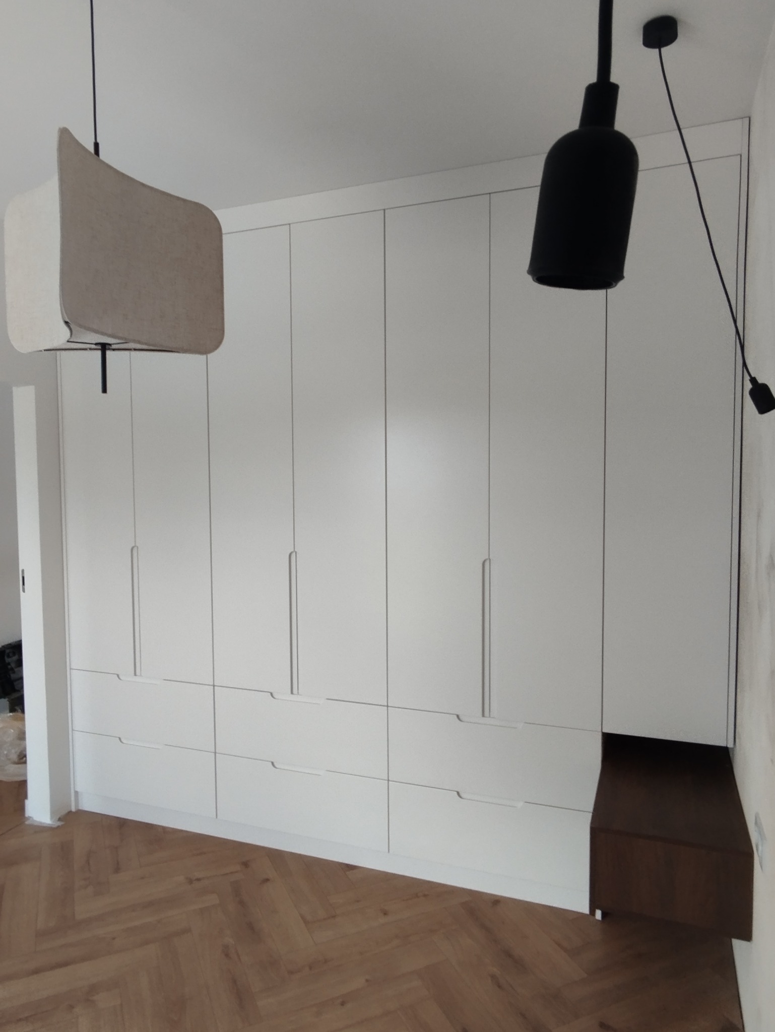 Biała szafa wnękowa z szufladami i minimalistycznymi uchwytami, oświetlona lampami wiszącymi w nowoczesnym wnętrzu z parkietem w jodełkę.