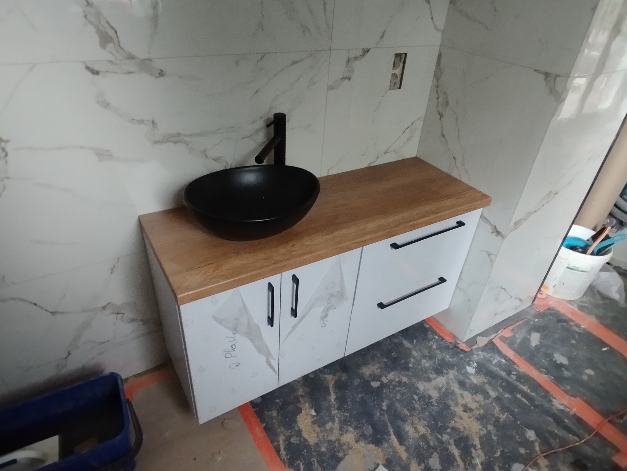 Nowoczesna szafka łazienkowa z czarną umywalką i drewnianym blatem w Radomsku. Białe fronty szafek z minimalistycznymi uchwytami. Marmurowe płytki na ścianie.