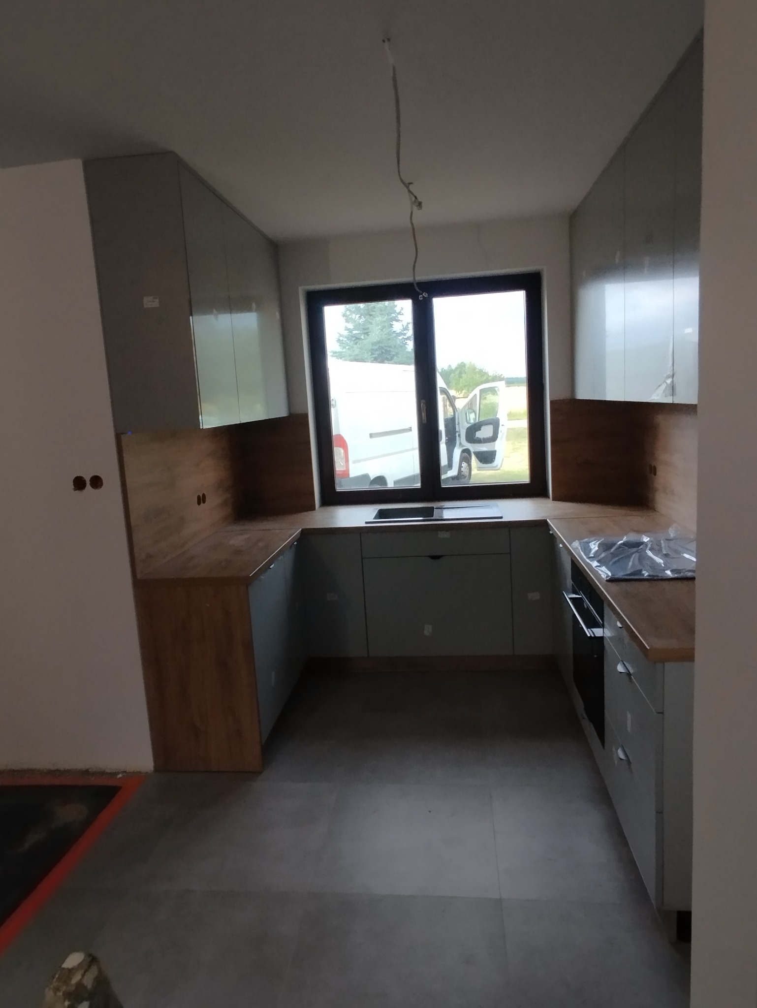 Jasna kuchnia w trakcie montażu: szare szafki, drewniany blat, widoczne niedokończone elementy instalacji elektrycznej. Przez okno widoczny samochód dostawczy.