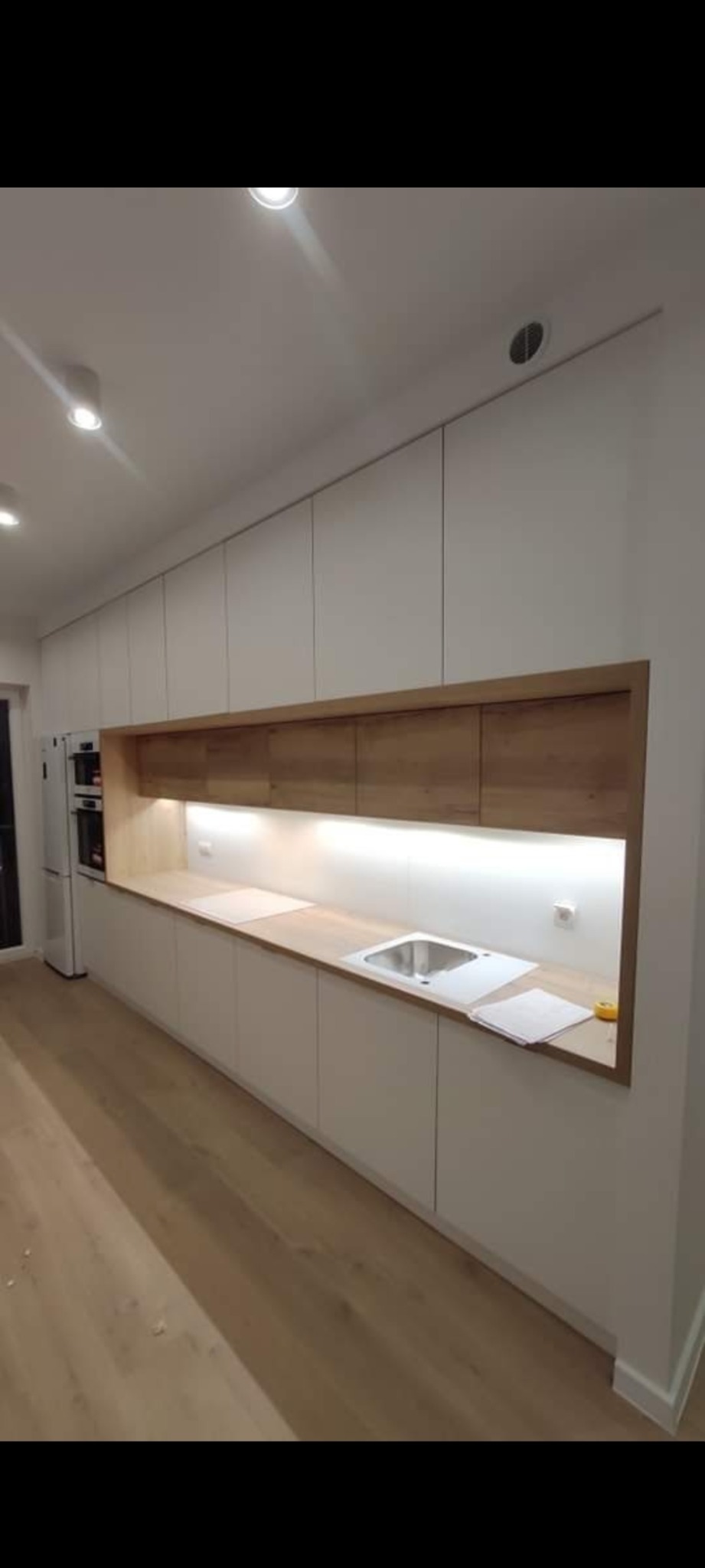 Nowoczesna kuchnia z białymi szafkami i drewnianymi akcentami. Długi blat roboczy z wbudowanym zlewem. Minimalistyczny design z oświetleniem LED pod szafkami.