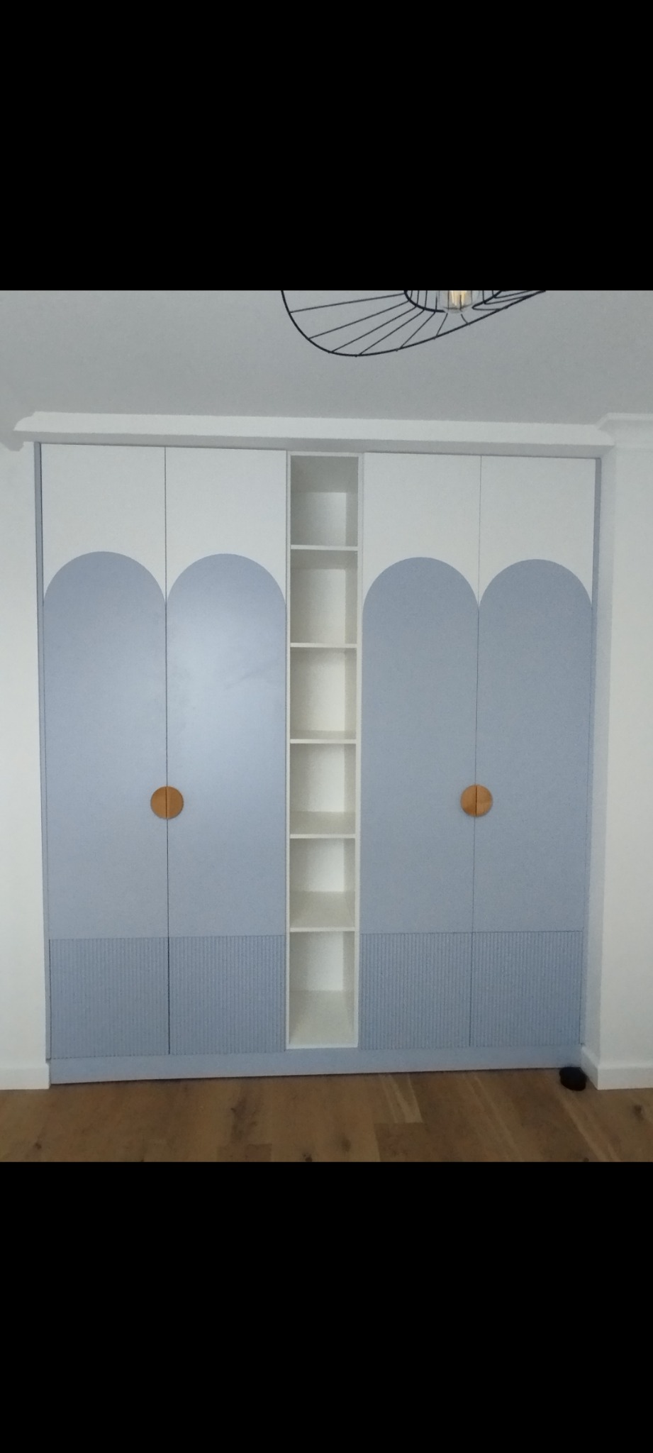 Biało-błękitna szafa z drewnianymi uchwytami i otwartą półką, na tle parkietu. Fronty zdobione motywem łuku i ryflowanym panelem dolnym. Nowoczesny design.