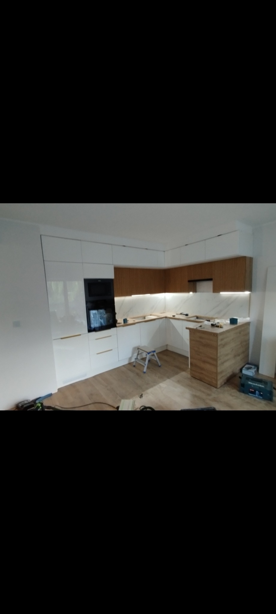 Nowoczesna kuchnia z białymi, lakierowanymi frontami i drewnianymi akcentami. Widoczny fragment wyspy kuchennej i oświetlenie LED pod szafkami górnymi. Na podłodze panele.