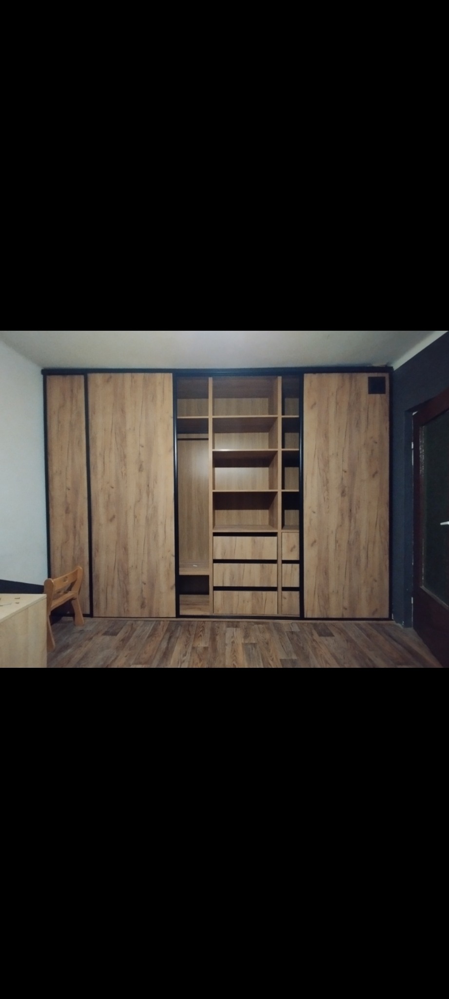 Duża szafa wnękowa z przesuwnymi drzwiami w odcieniach drewna, z czarnymi obramowaniami, na tle drewnianej podłogi. Widoczny fragment krzesła.