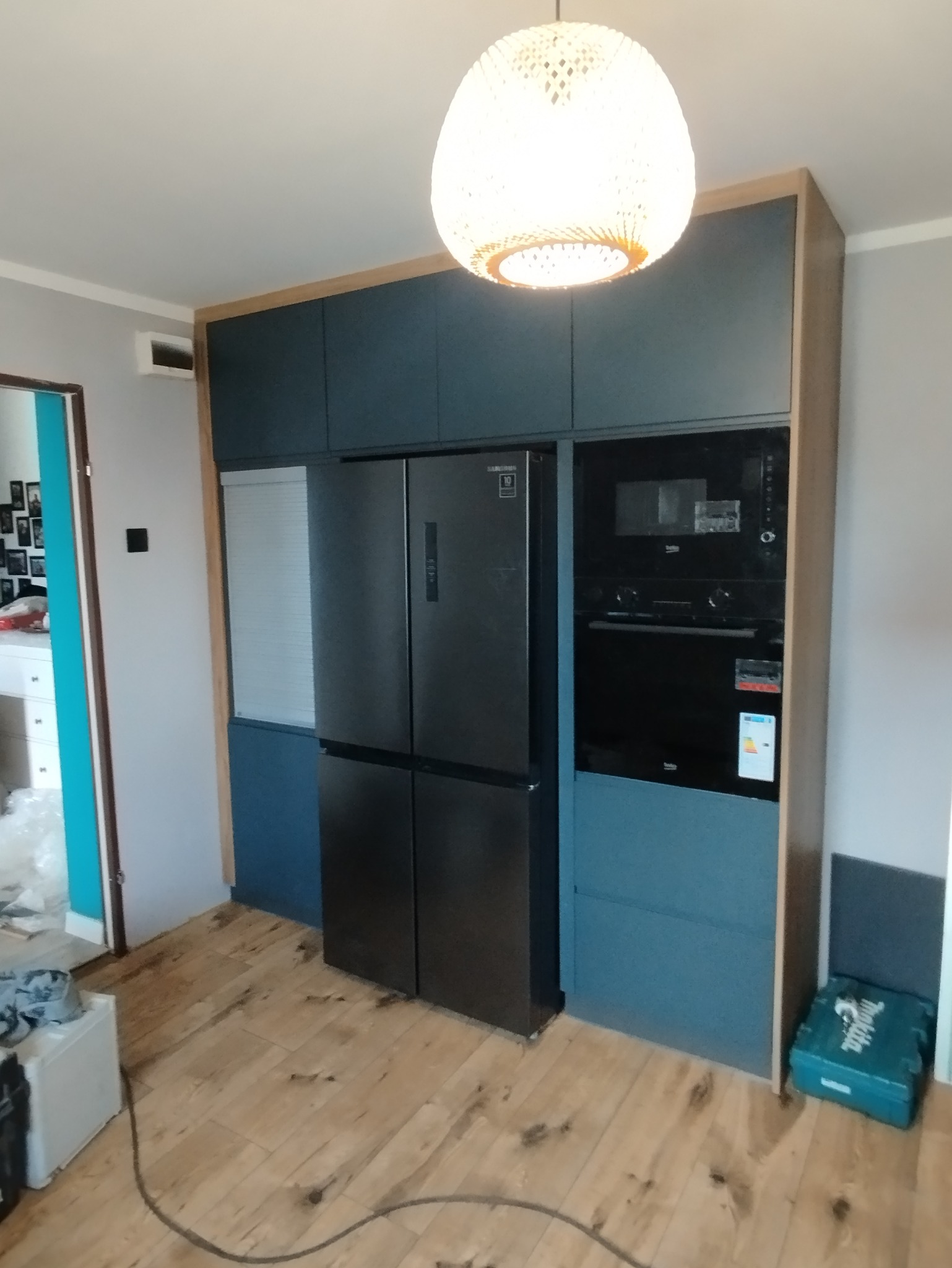 Nowoczesna zabudowa kuchenna w odcieniach szarości i drewna, z wbudowaną lodówką side-by-side i piekarnikiem. Na podłodze drewniane panele, a w tle widoczny fragment innego pomieszczenia.