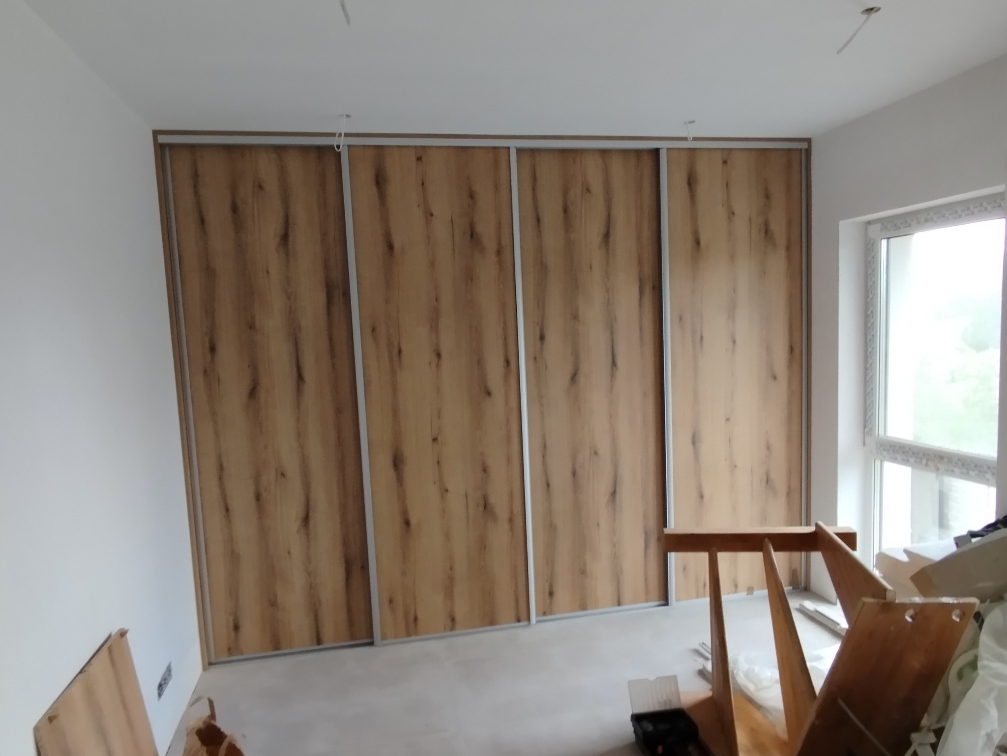 Zamontowana szafa wnękowa z przesuwnymi drzwiami o drewnianej fakturze, widoczna w surowym wnętrzu z oknem i elementami montażowymi.