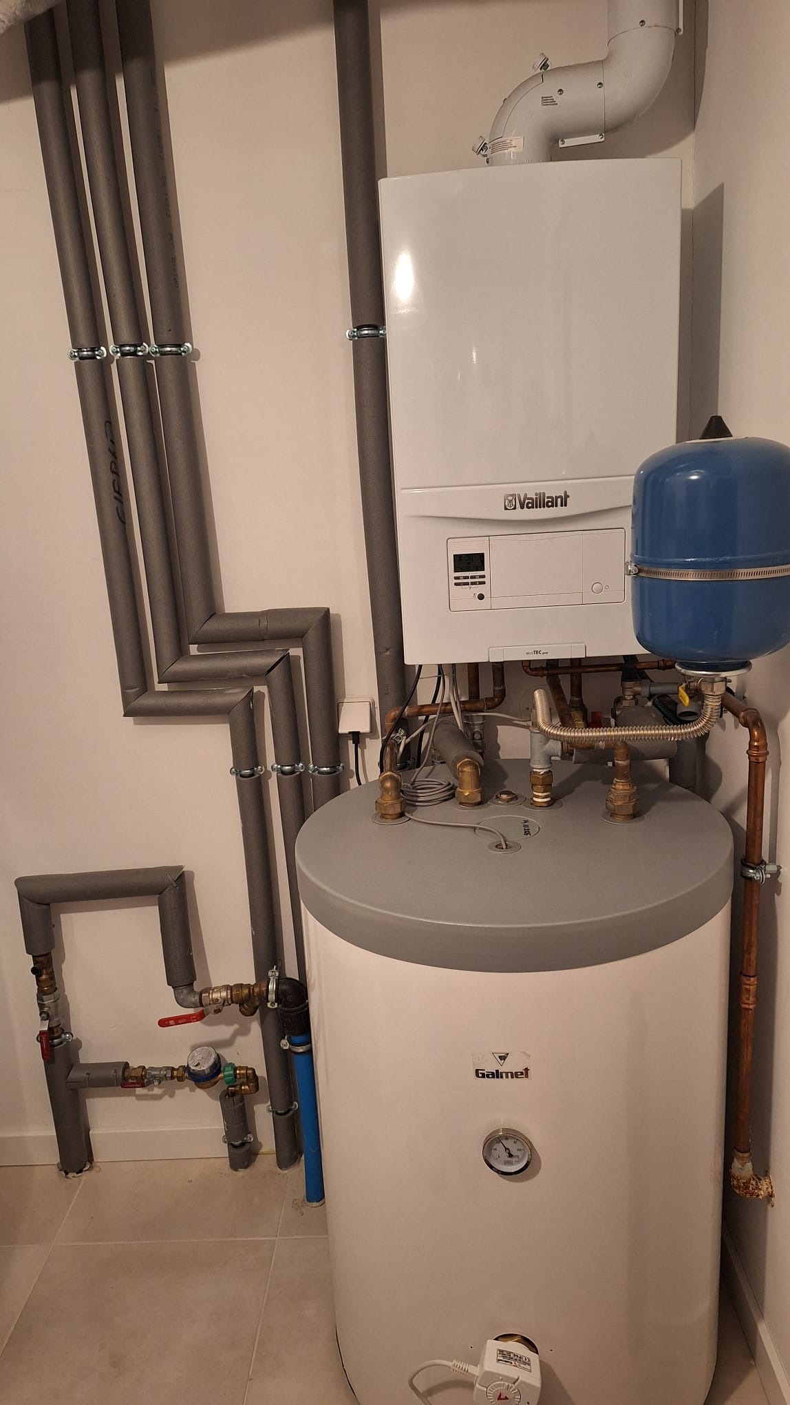 Biały piec gazowy Vaillant z niebieskim zbiornikiem, zamontowany nad białym bojlerem Galmet. Rury w szarej izolacji prowadzone wzdłuż ściany.