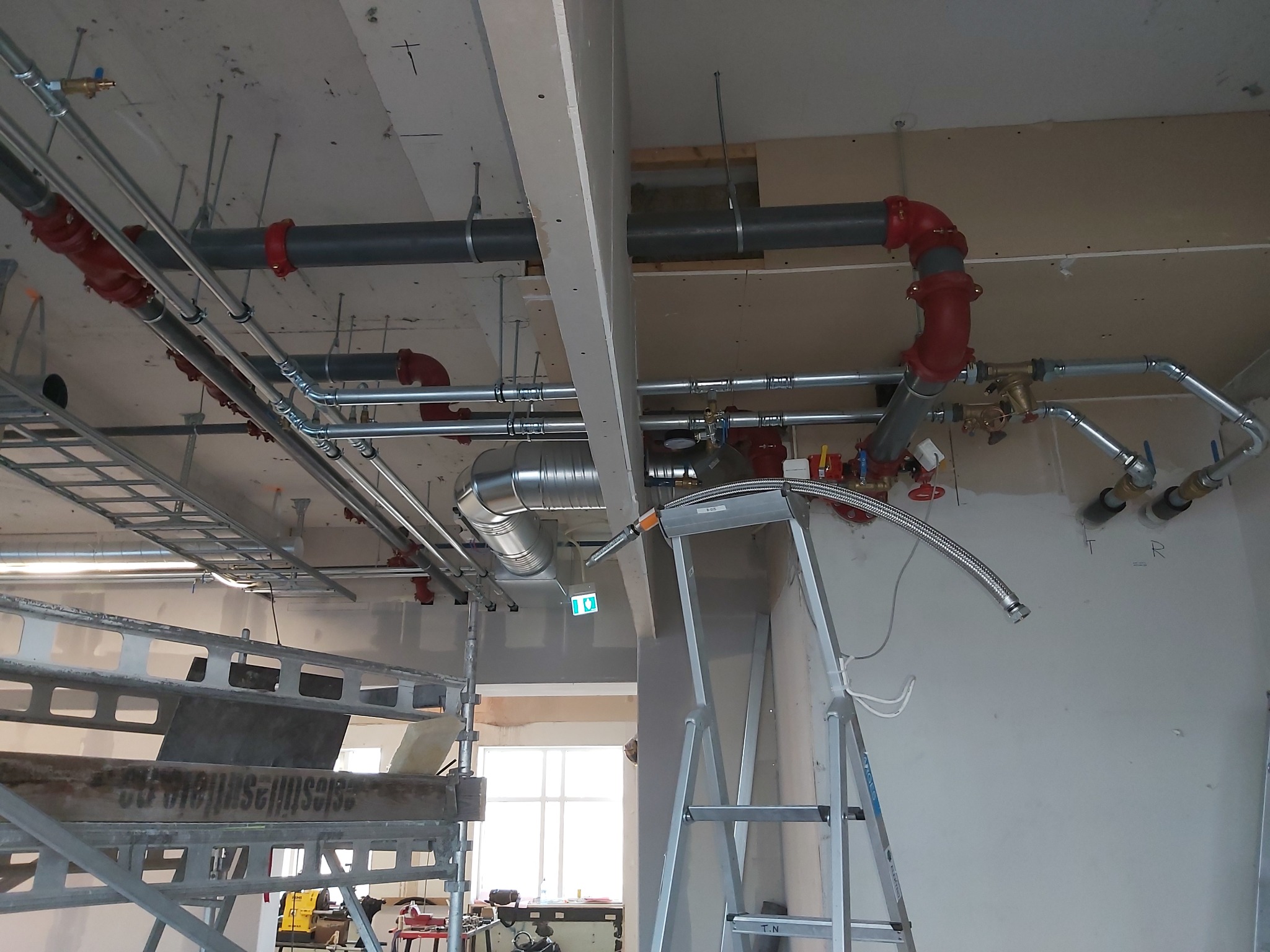 Instalacja rur pod sufitem w budynku w trakcie budowy. Widoczne rury w kolorze szarym i czerwonym, drabina i rusztowanie. Oświetlenie sufitowe i okno w tle.