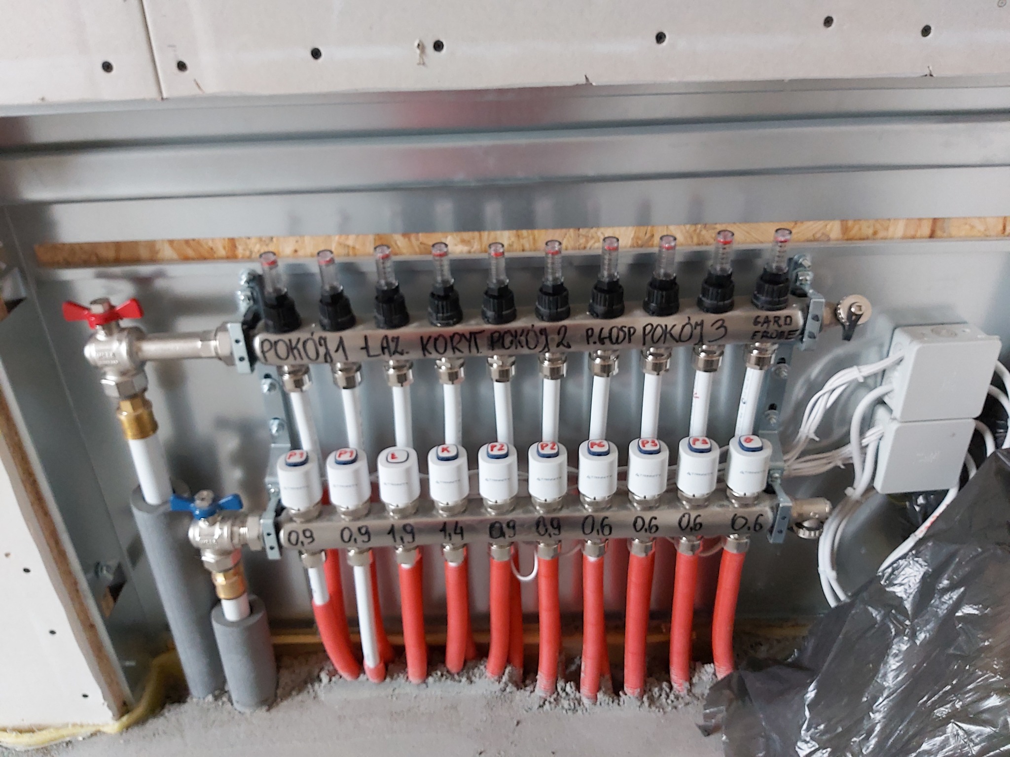 Rozdzielacz ogrzewania podłogowego z rurami w kolorze czerwonym i białym, opisany dla poszczególnych pomieszczeń, zamontowany na ścianie w surowym stanie.