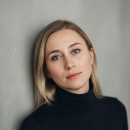 Kancelaria Adwokacka Adwokat Marta Bortel-Borowska - Portret kobiety o jasnych włosach i niebieskich oczach, opierającej się o szarą ścianę. Ubrana w czarny golf, patrzy w obiektyw z delikatnym uśmiechem.