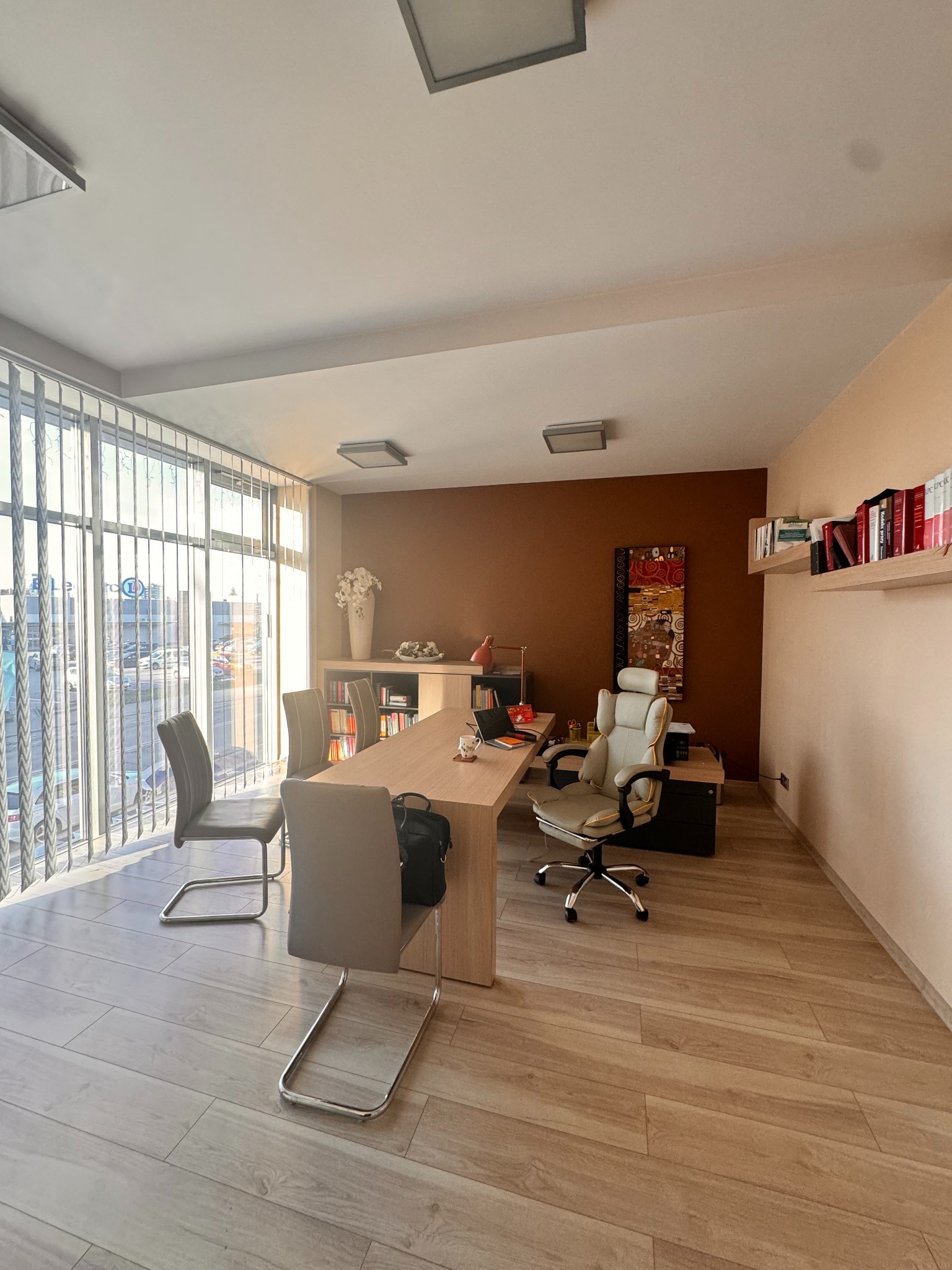 Jasne biuro adwokackie z dużym oknem, biurkiem, krzesłami dla klientów i regałem z książkami prawniczymi. Stonowana kolorystyka, nowoczesny design.