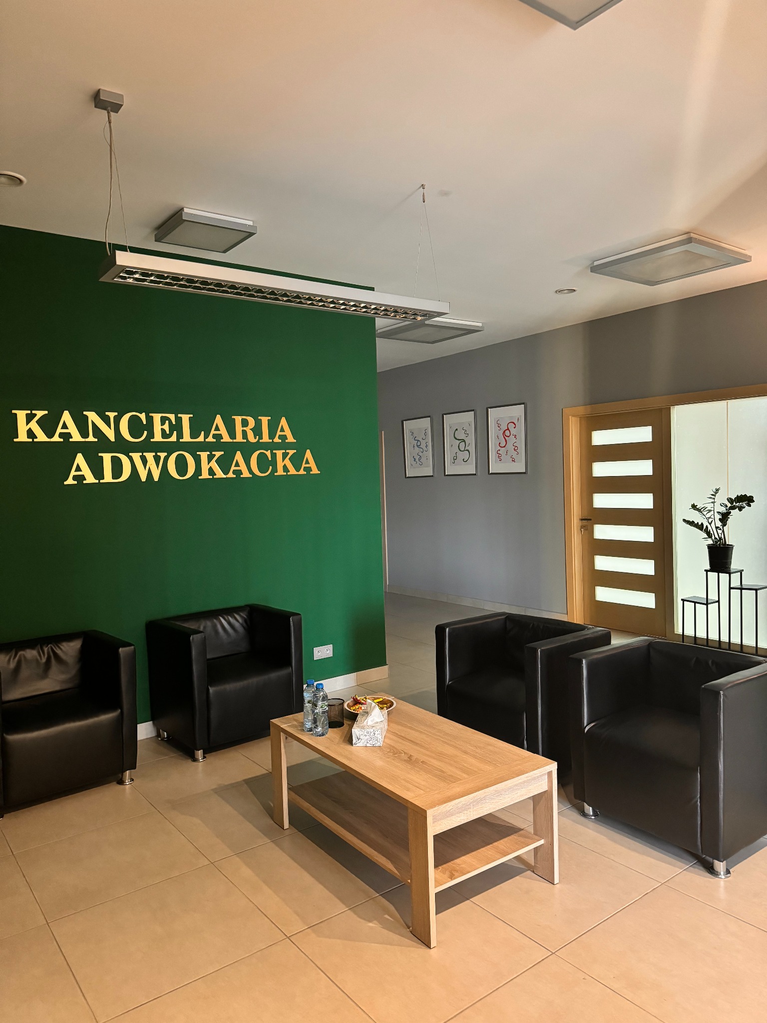 Wnętrze kancelarii adwokackiej: zielona ściana z napisem 'Kancelaria Adwokacka', czarne fotele, drewniany stolik kawowy, grafiki na ścianie i roślina doniczkowa.