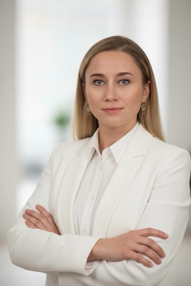 Portret kobiety w białym garniturze, z blond włosami i niebieskimi oczami, skrzyżowane ręce, delikatny uśmiech, jasne tło, profesjonalny wizerunek.